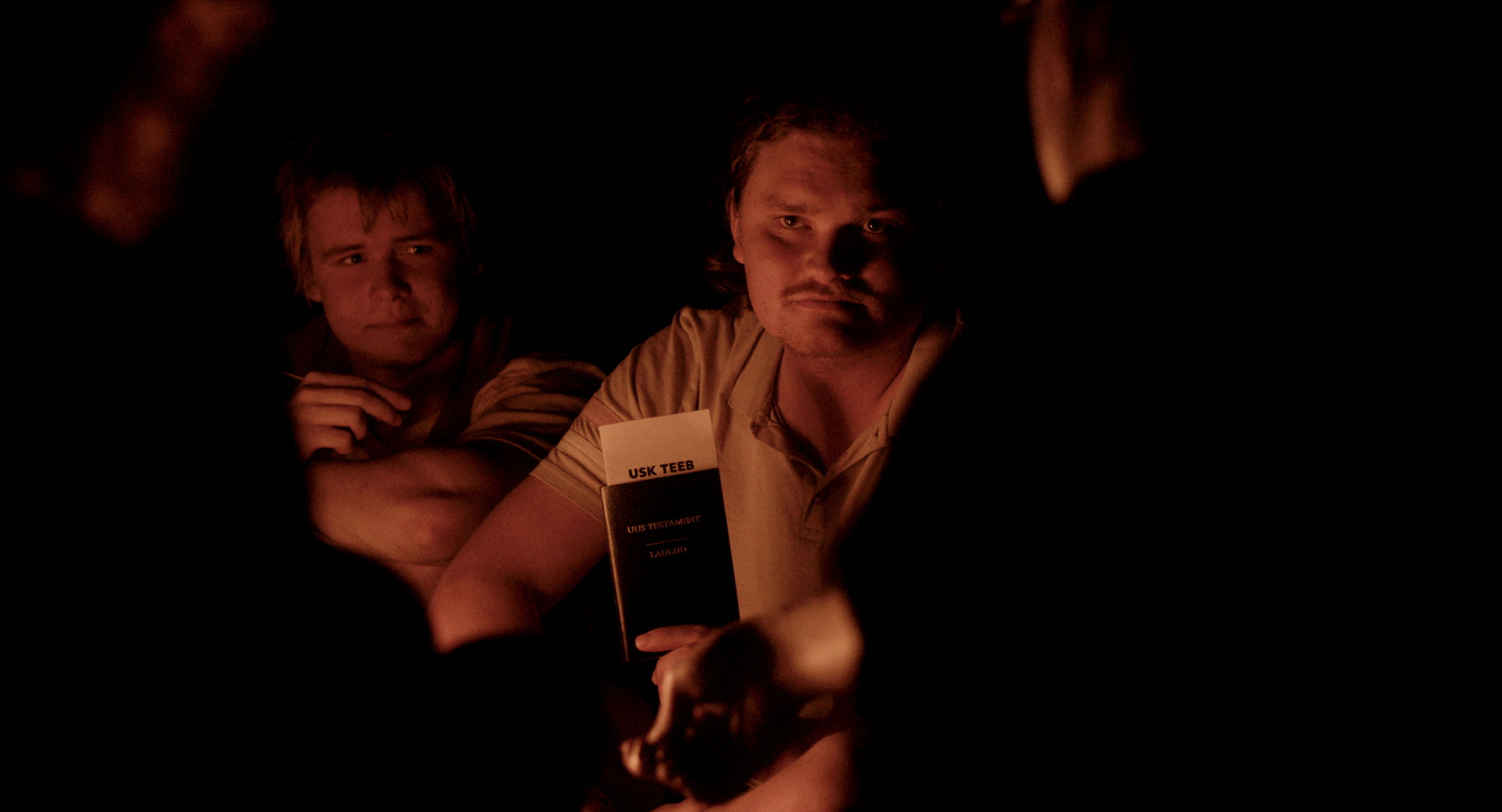 People in a dimly lit environment, possibly around a fire, with one person holding a booklet, looking intently at something in front of them.