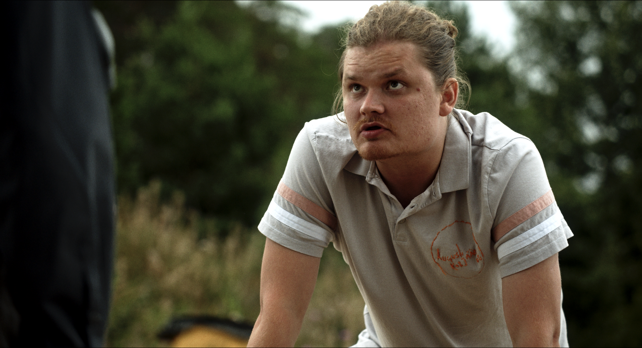 A young man with light skin, curly hair, and a goatee, wearing a beige polo shirt, appears to be in an outdoor setting surrounded by greenery. He is looking upward with a serious expression and leaning forward.