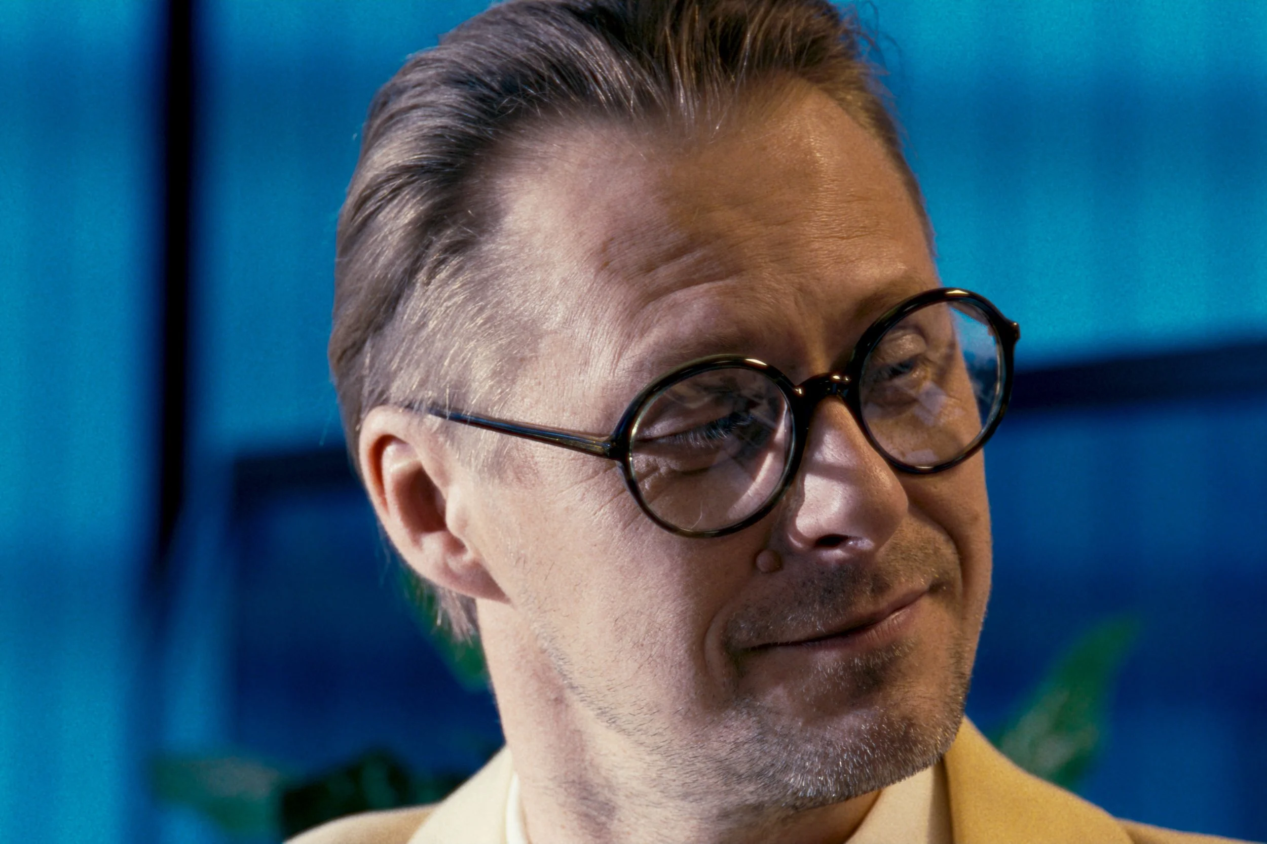 Close-up of a man with glasses and gray hair, wearing a yellow shirt, against a blue background.