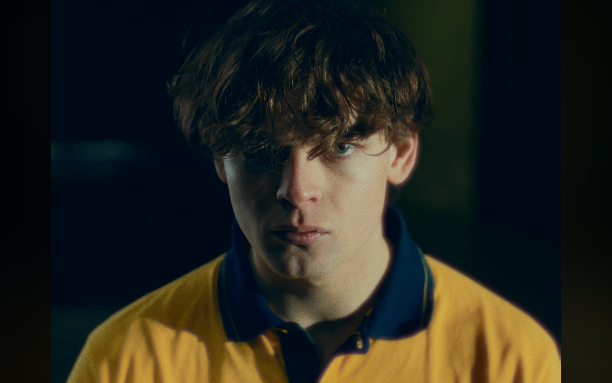 Close-up of a young person with curly brown hair and blue eyes, wearing a yellow jacket and navy blue shirt, looking directly at the camera with a serious expression.