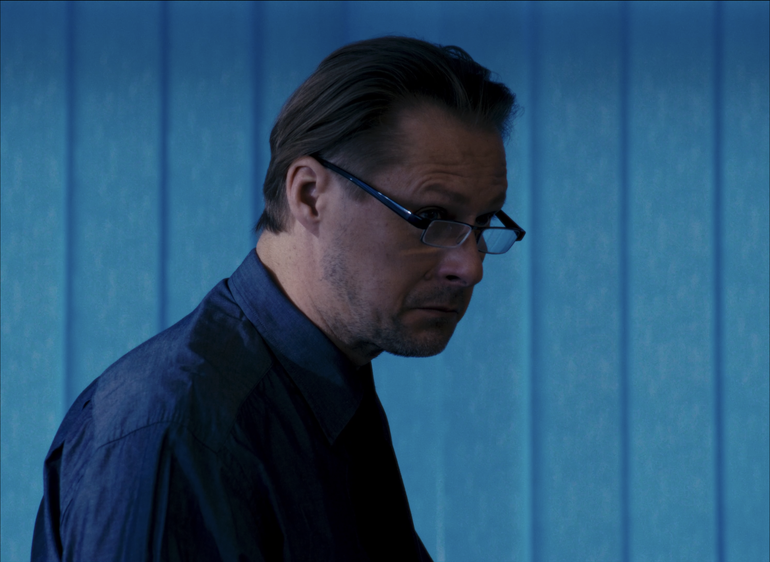 A man with dark hair, wearing glasses and a dark shirt, looking downward against a blue vertical panel background.