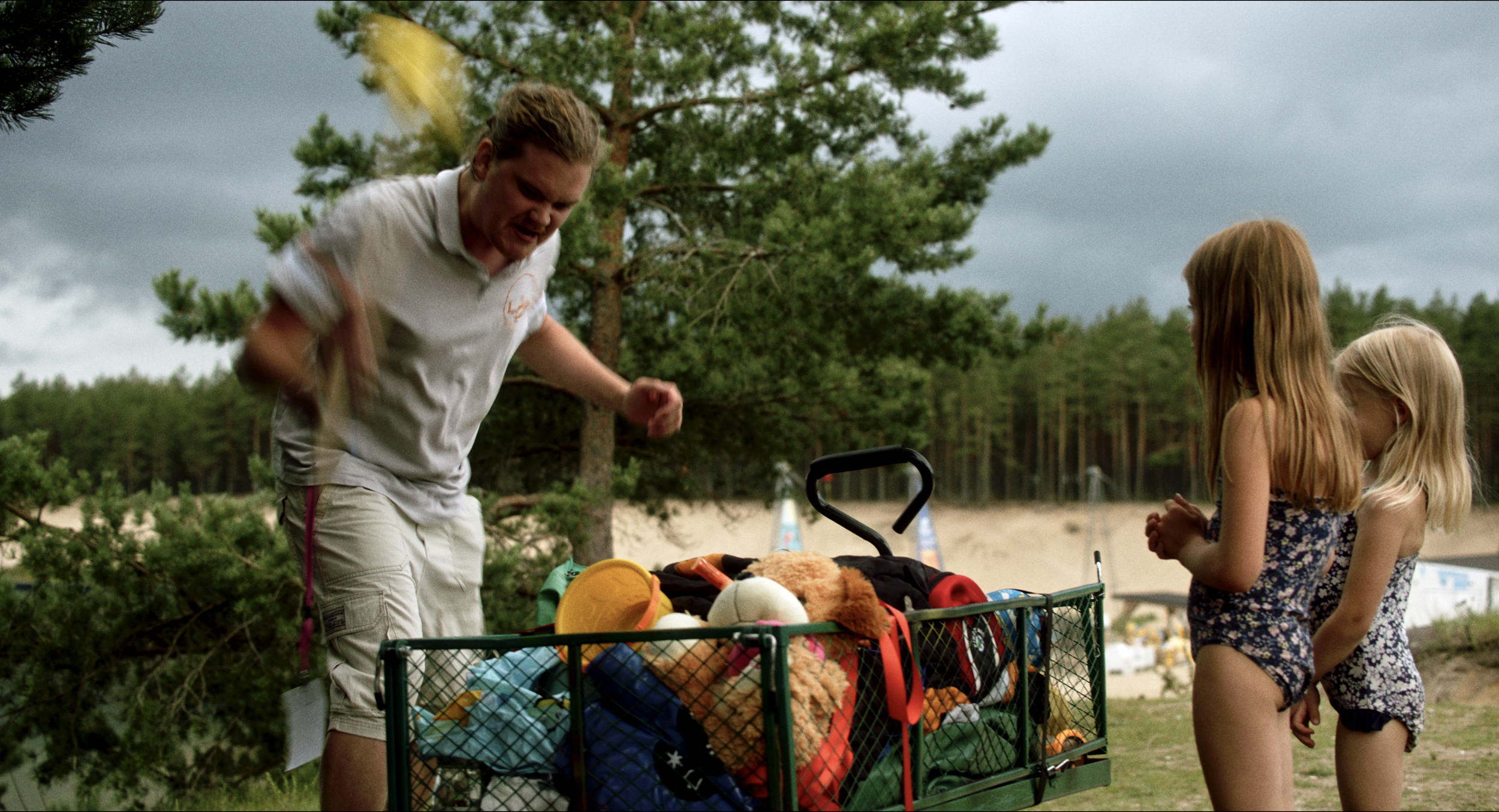 A man with long hair and a beard wearing a white polo shirt and khaki shorts pushing a shopping cart filled with toys and stuffed animals. Two young girls with long blonde hair, one with a floral dress and the other with a patterned shirt, standing n