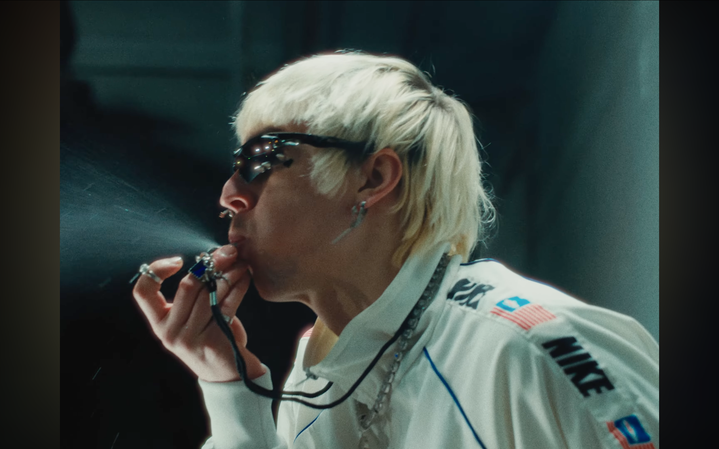 A person with platinum blonde hair wearing sunglasses, multiple rings, and ear jewelry, dressed in a racing suit with sponsor logos, sitting with a hand near their lips in a dark setting.