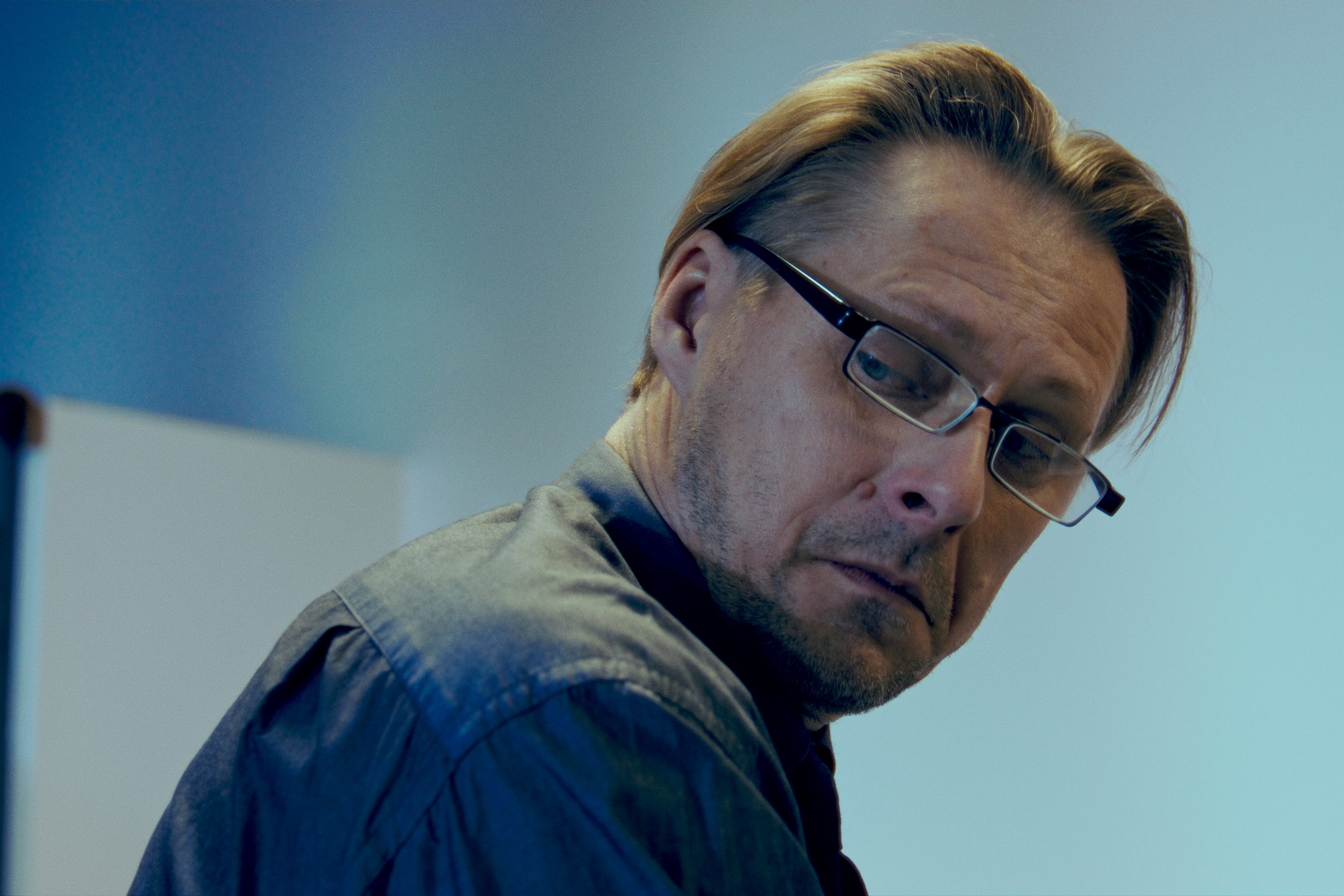 A man with blond hair, glasses, and a gray shirt is looking down, with a serious expression in a softly lit room with a blue background.