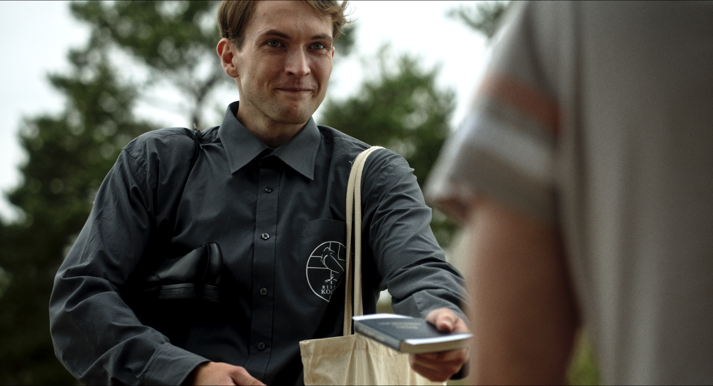 A young man with light skin, short light brown hair, wearing a gray button-up shirt with a logo, is smiling slightly and holding a smartphone. He is outdoors with blurred trees in the background.