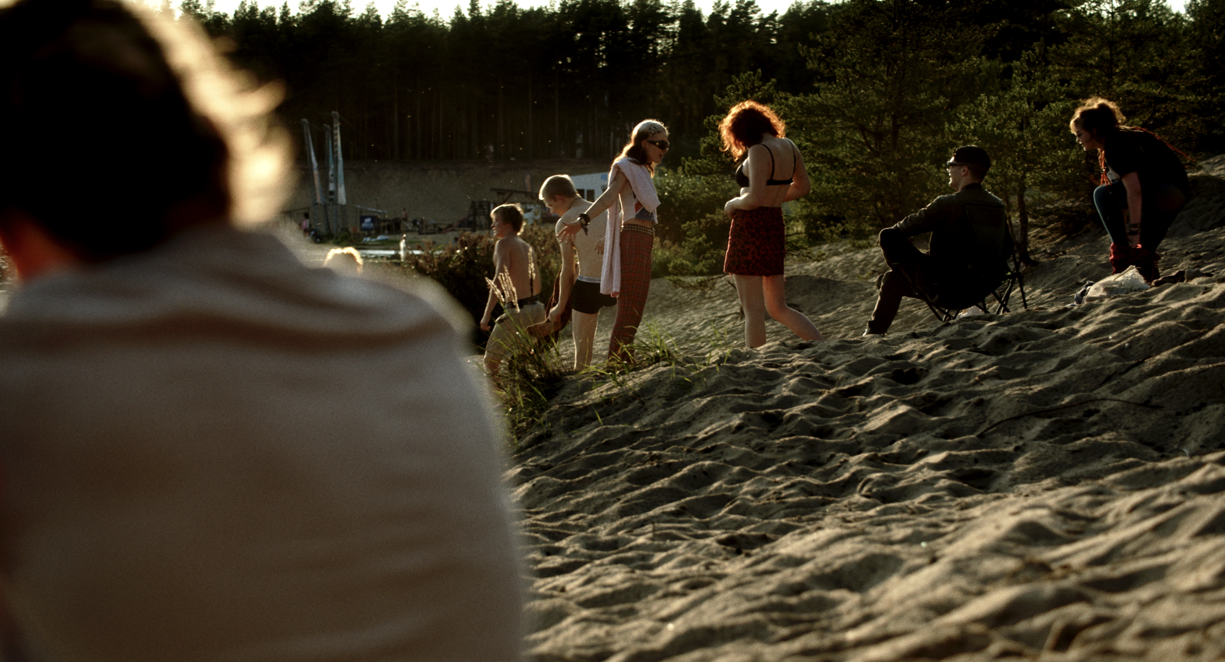 People gathered on a sandy hill at sunset, with some sitting and others standing, near trees and structures in the background.