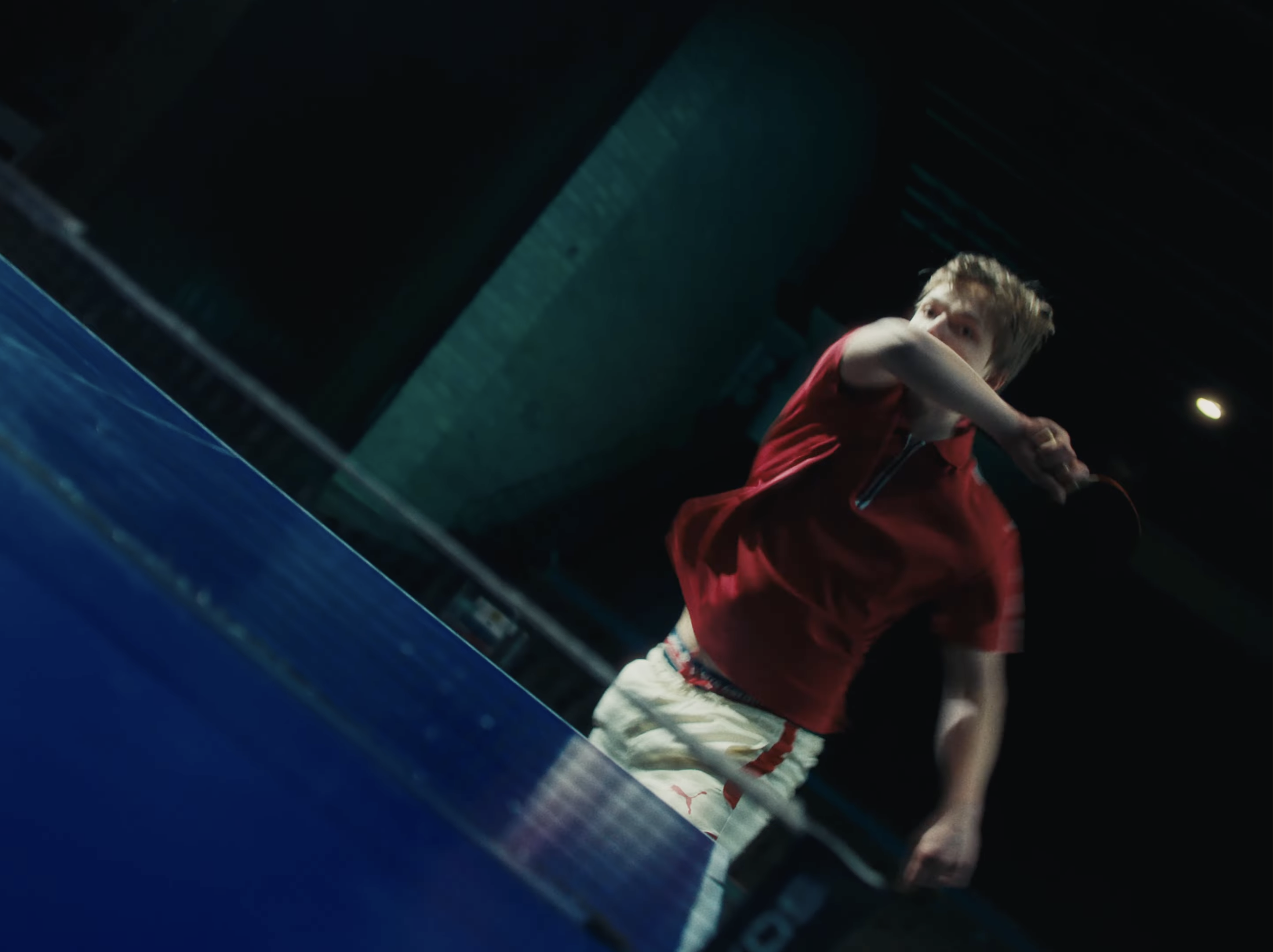 A young boy with messy blonde hair wearing a red shirt and white shorts, possibly playing a sport indoors with a blue surface.