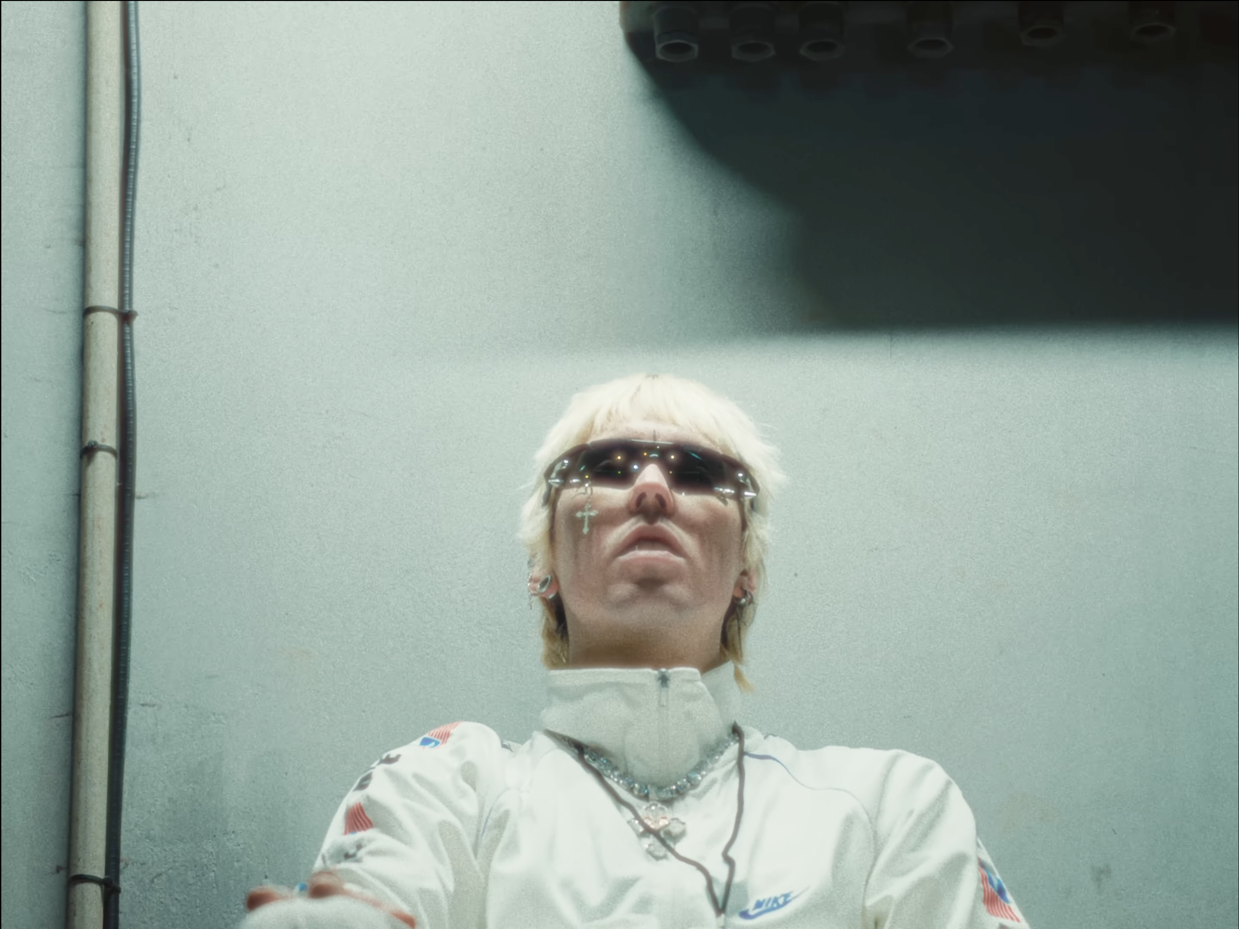 A person with short blonde hair wearing dark sunglasses, a white Nike jacket with red and blue accents, a beaded necklace, and earrings standing against a gray wall with visible pipes.