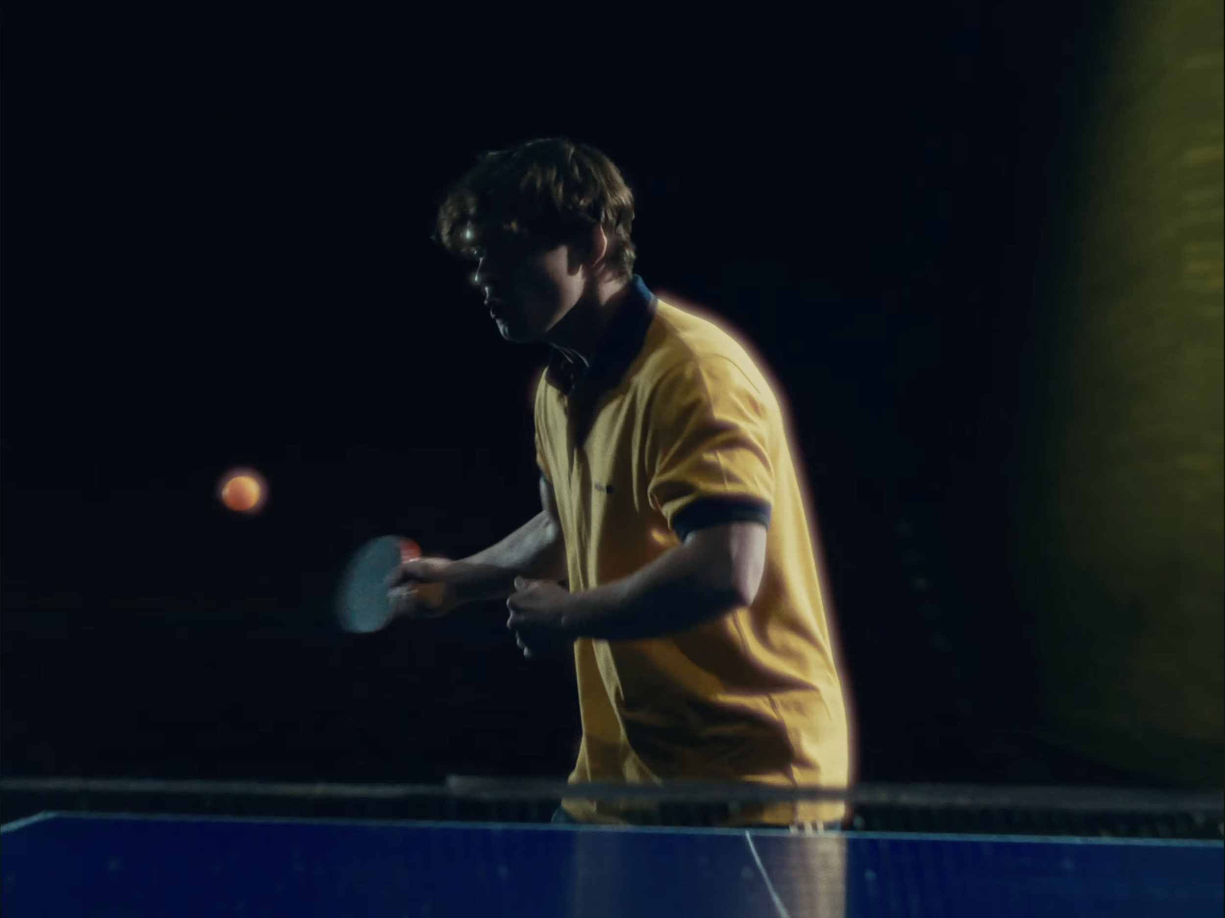 A young person with curly hair wearing glasses and a yellow shirt playing table tennis at night, illuminated by artificial lighting.