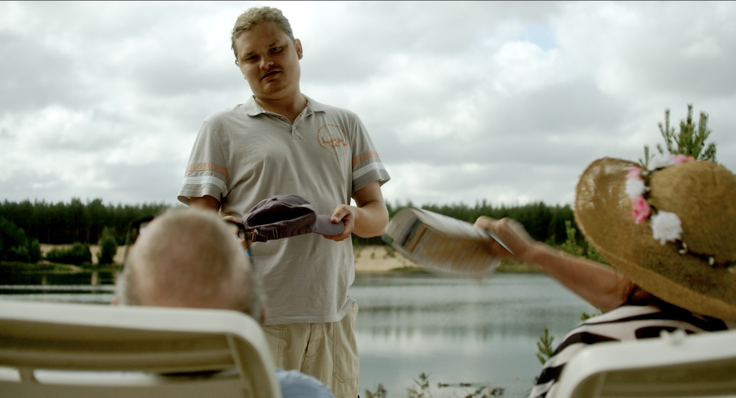 A young man in a light-colored polo shirt and beige shorts is standing outdoors by a body of water, holding a cap and some papers, while a woman with a straw hat decorated with flowers extends her hand holding a flyer or brochure toward him. Other pe
