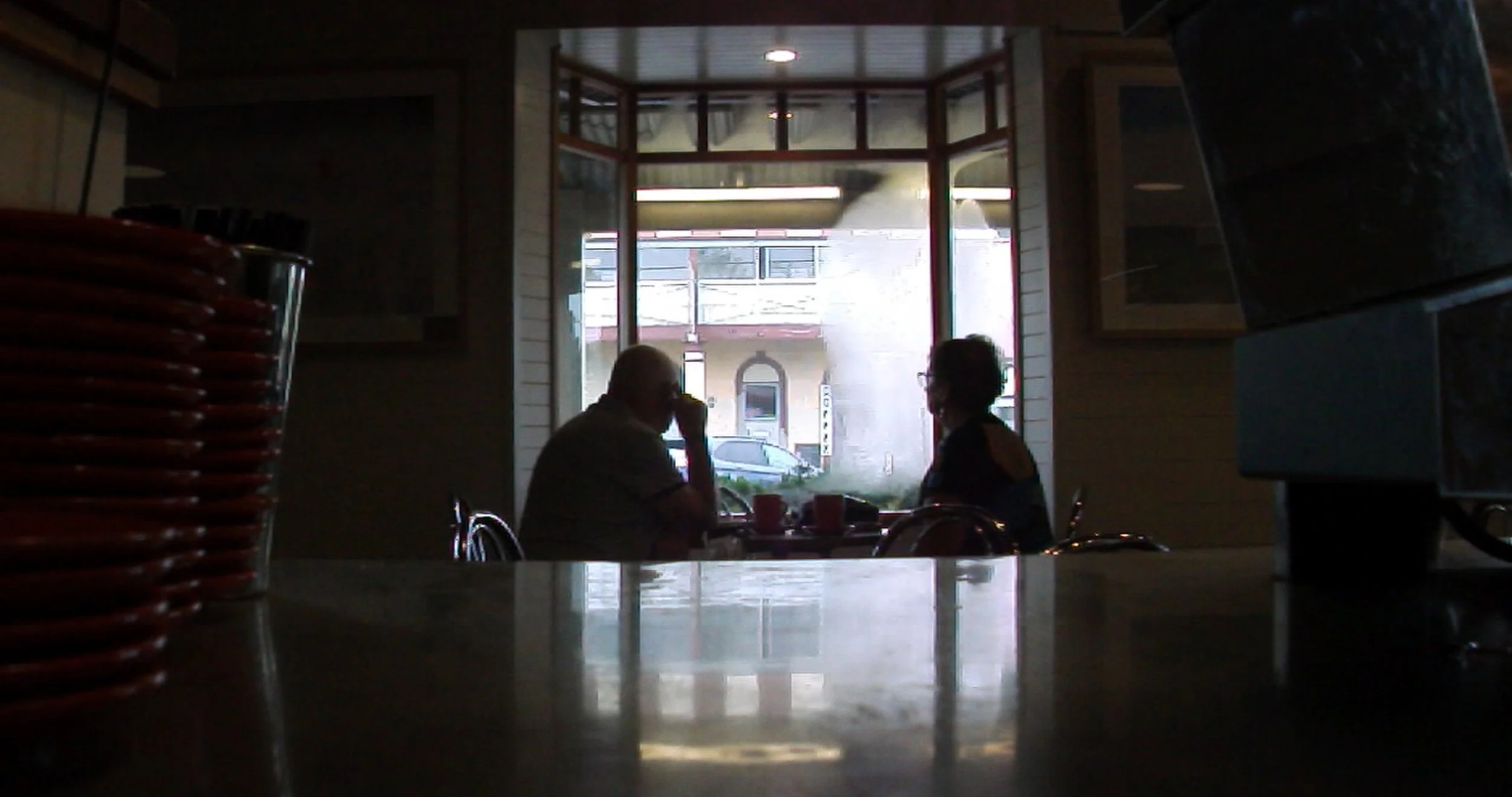 Two people sitting at a table in a darkly lit restaurant, with a window showing the street outside.