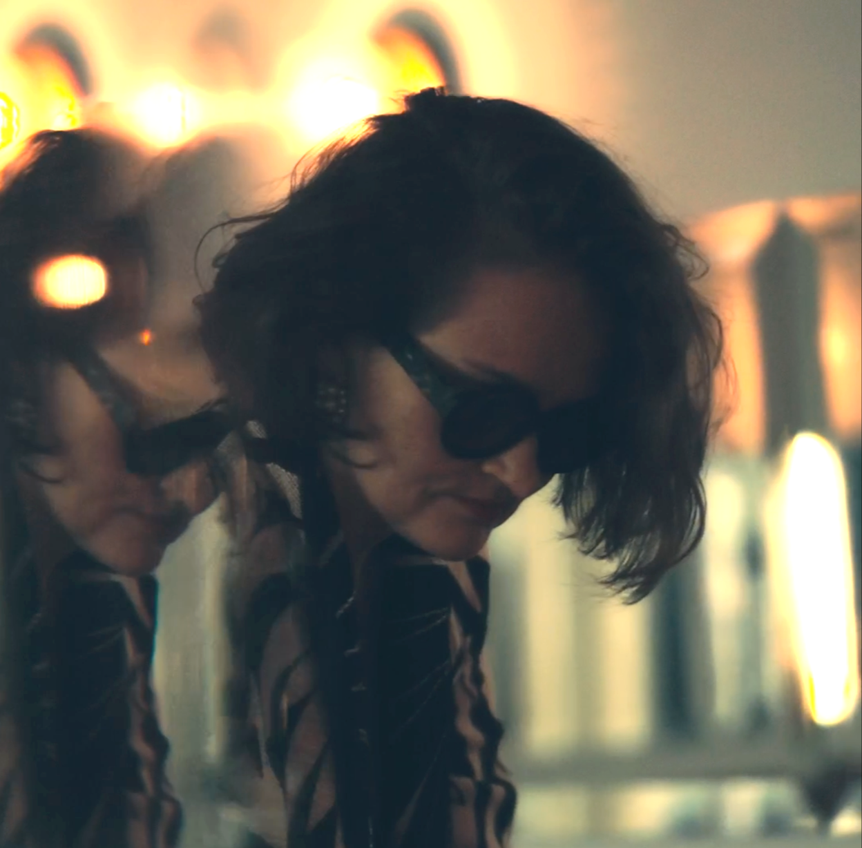 Two women with dark wavy hair wearing sunglasses and striped shirts, looking down, with a blurred background featuring bright lights.