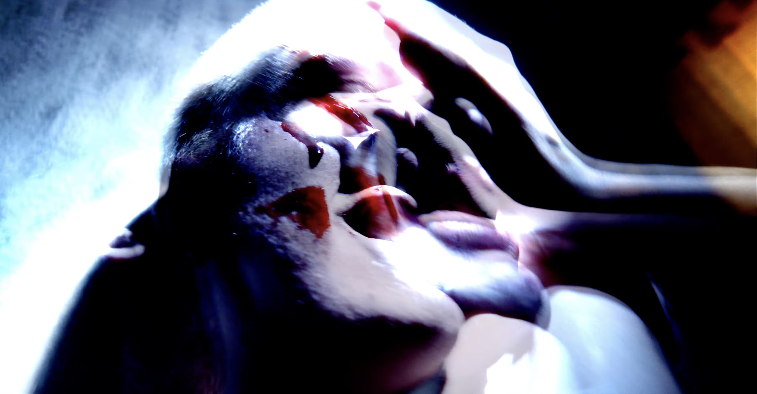 A close-up of a person's face with makeup or face paint, eyes closed, being touched on the forehead and cheeks by hands with red paint or blood on them, in a dramatic and intense scene with dark and vibrant lighting.