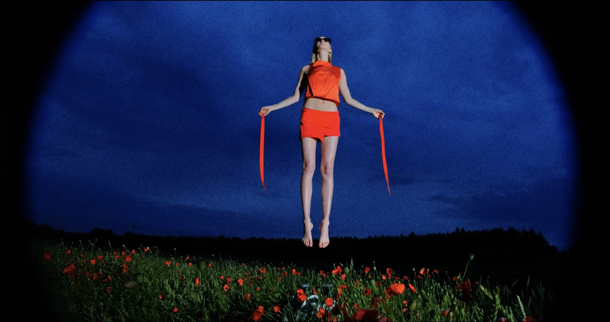 A person with long hair wearing sunglasses and a red outfit with long red ribbons is floating in the air above a field of red flowers, against a dark blue sky.