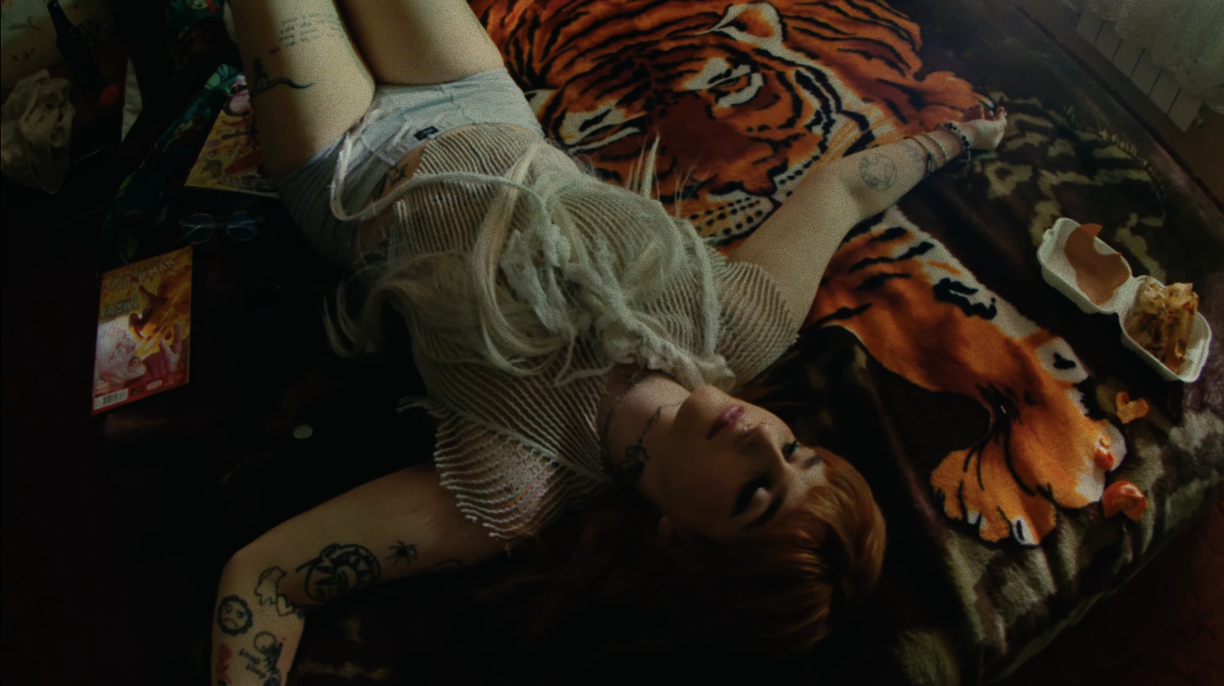 A person lying on a tiger-patterned blanket, with a mesh top and tattoos on their arm. There are snacks and a magazine around them, on a dark surface.
