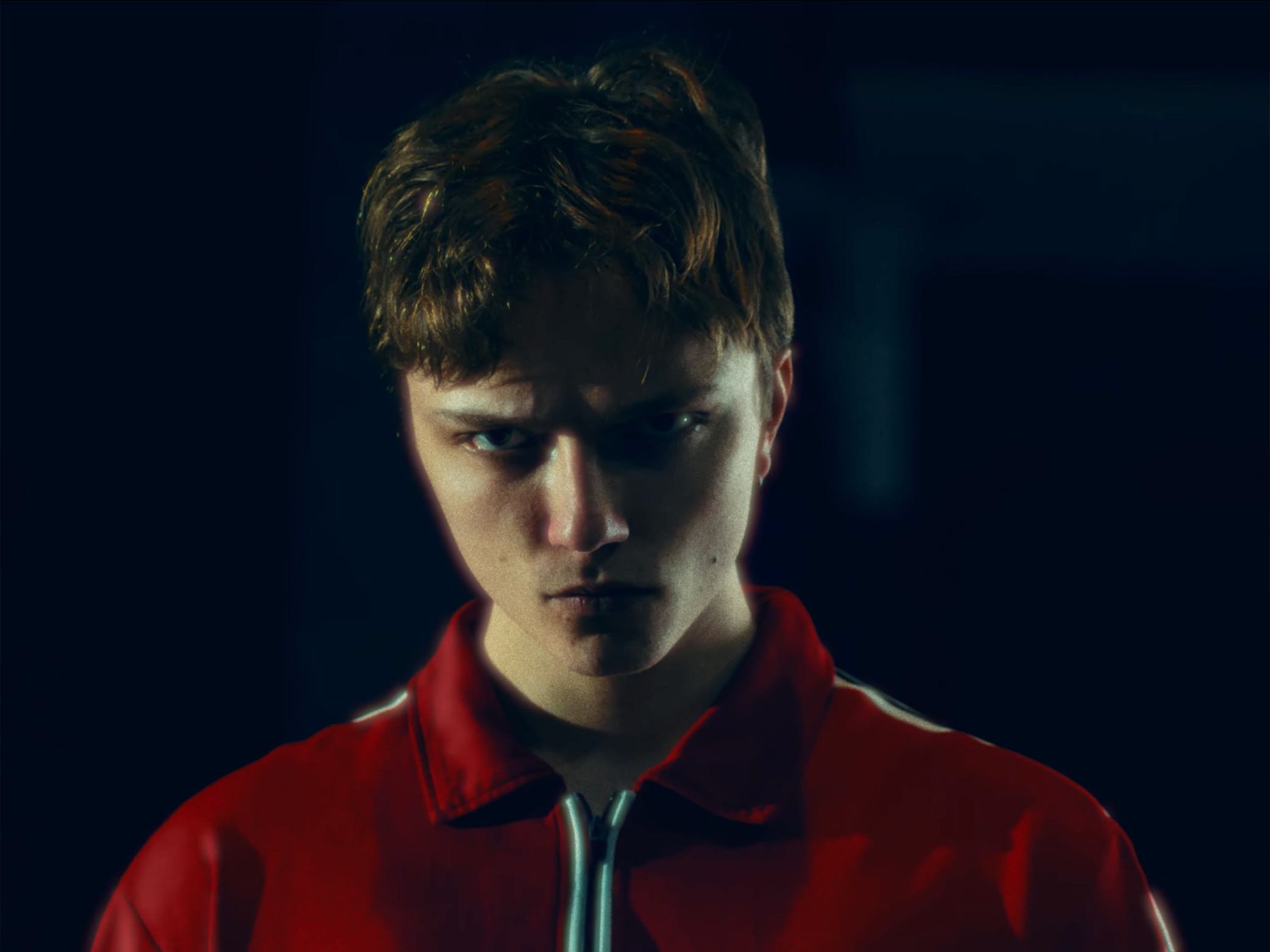 A close-up image of a young man with light skin, brown hair, and intense eyes, wearing a red jacket with a zipper, against a dark background.
