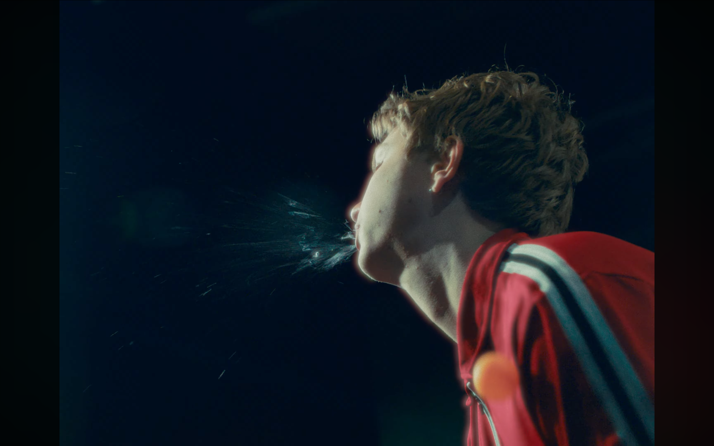 A person with brown hair wearing a red jacket with white stripes on the sleeves, in profile, appears to be spitting or coughing, with water and mist visible.