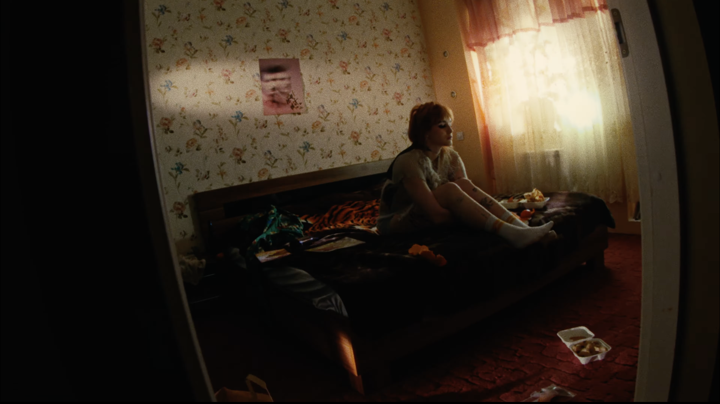 The image shows a woman with red hair sitting on a bed in a room with floral wallpaper. She is wearing a light-colored shirt and socks, and appears to be looking thoughtfully out the window. There are various items on the bed and a food container on 