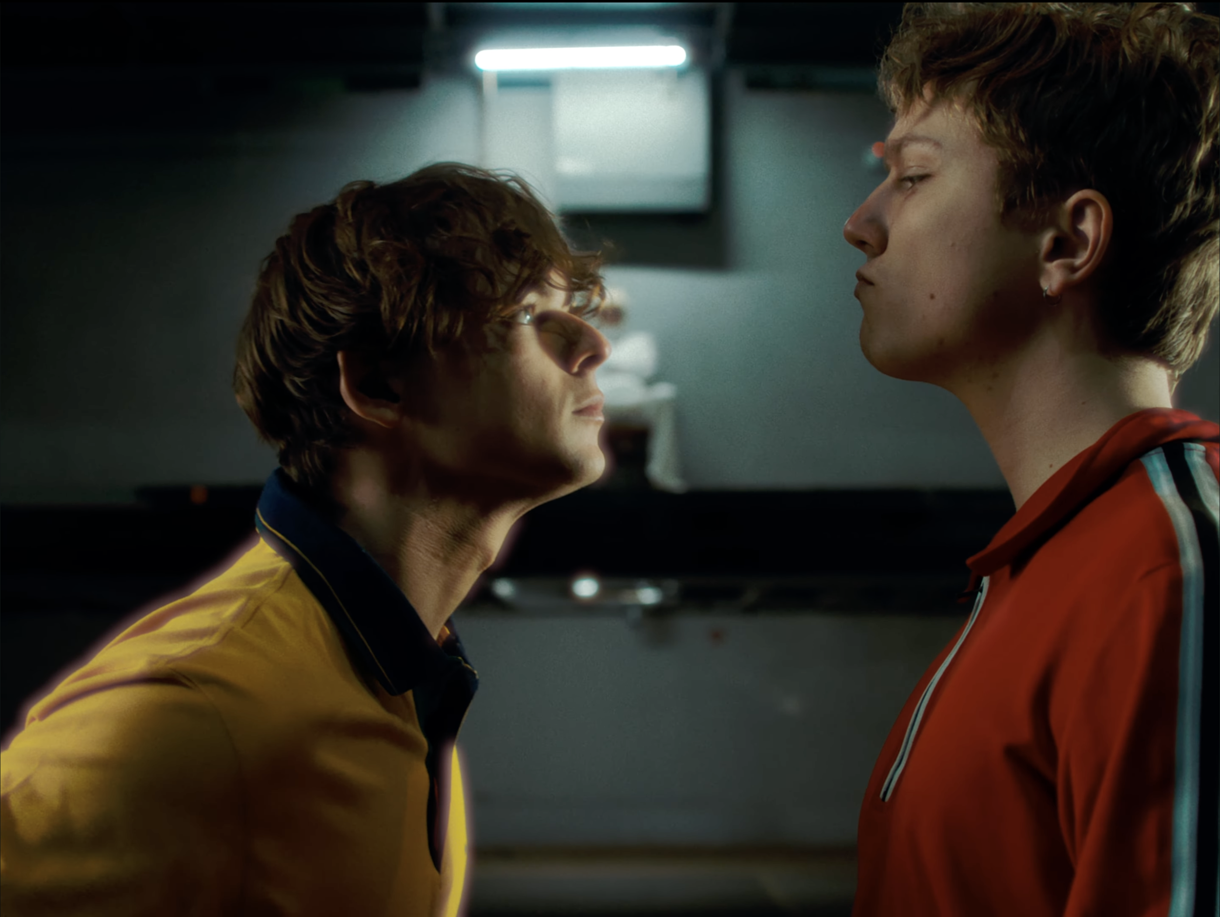 Two young men face each other closely in a dimly lit room, one wearing a yellow sports jersey and the other in a red sports jacket, with a blurred background and bright light overhead.