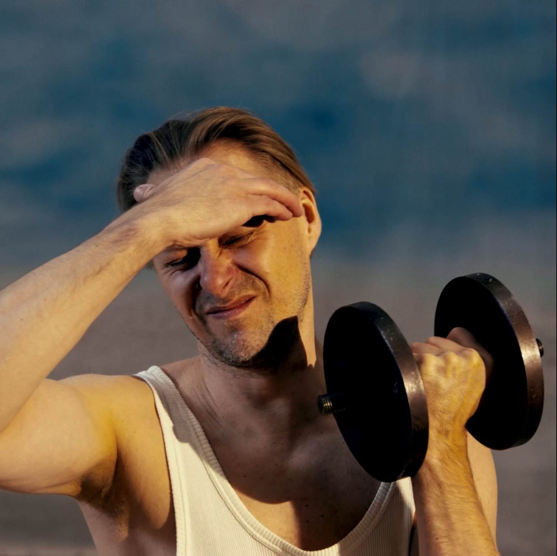 Young man lifting dumbbells at the gym, wearing a white tank top, with a cloudy sky background.