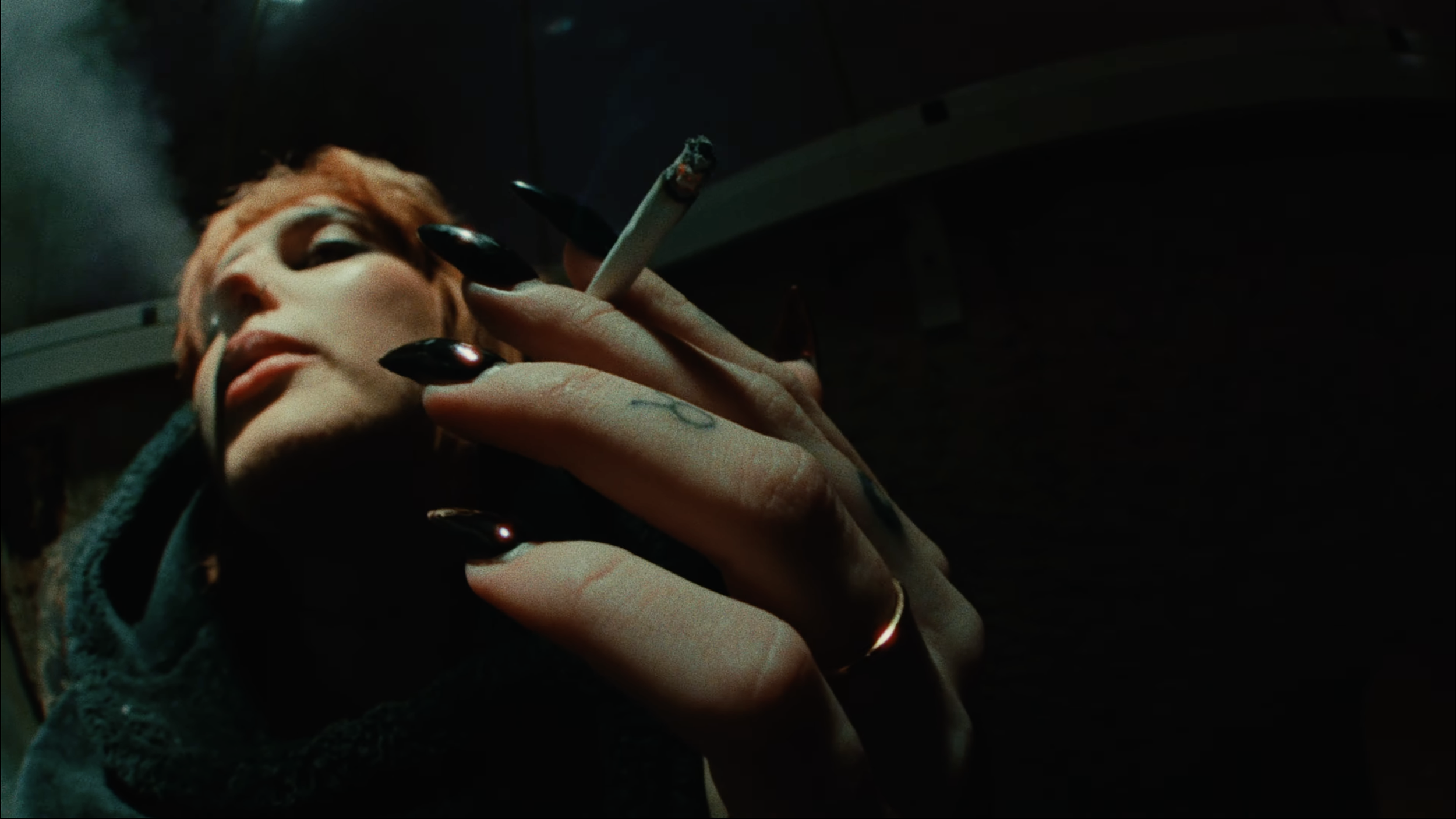 Close-up of a person's face and hand with long black nails, holding a cigarette in a dark setting.