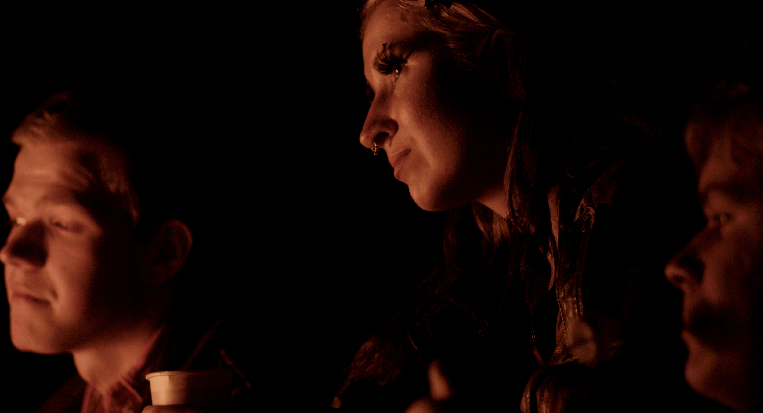 A woman with light skin and tattoos leaning over a table, surrounded by several young men, in a dark room illuminated by dim, warm lighting.
