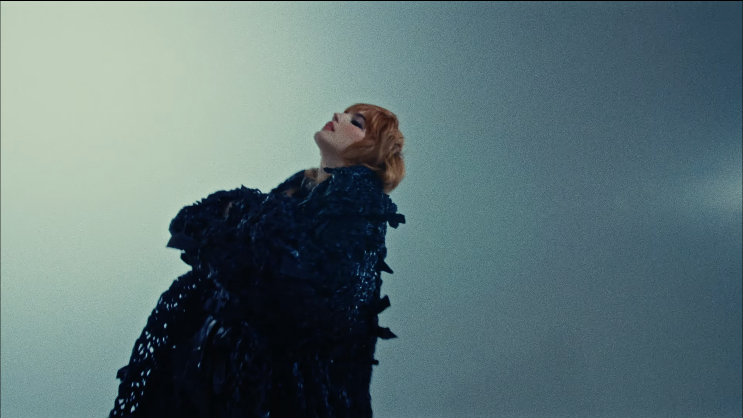 A woman with short, reddish hair and fair skin looking upward, wearing a dark, textured coat, standing against a gradient background of pale green to blue.