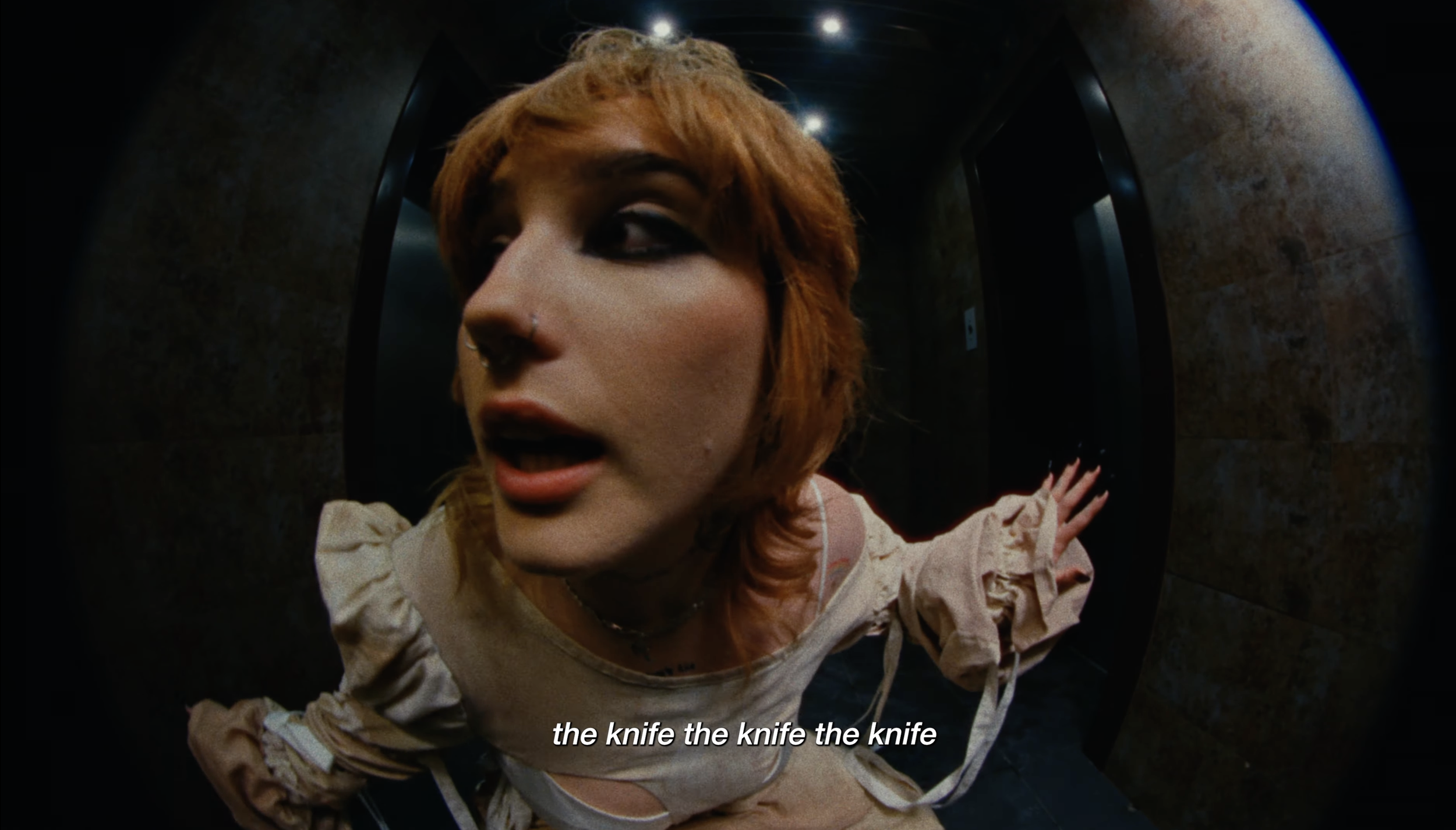 A young woman with short, reddish hair and makeup, wearing a beige top, is inside an elevator with dark walls. She appears to be looking out for something, and the scene is dark. The image has subtitles reading "the knife the knife the knife."