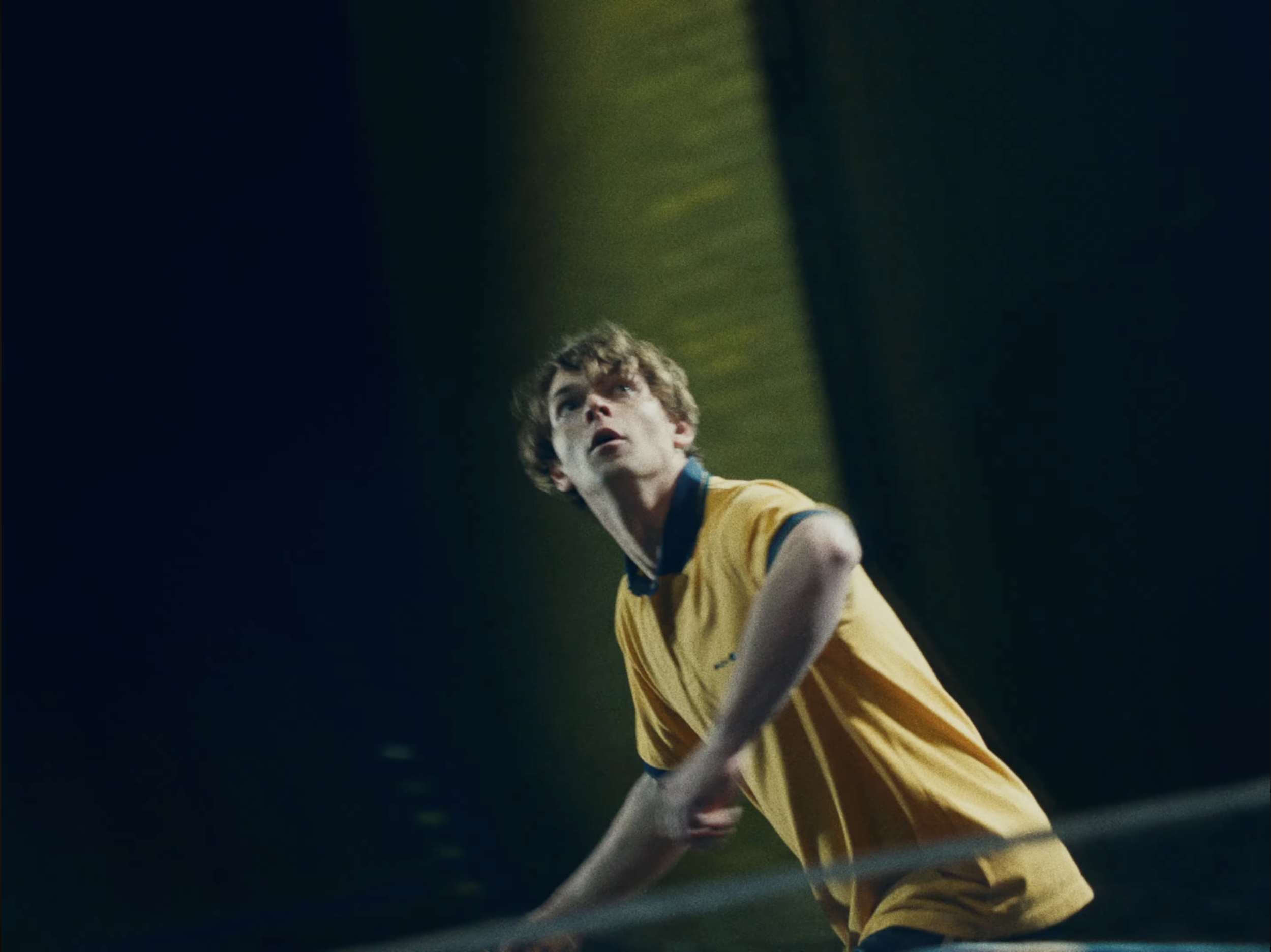 A young male soccer player in yellow uniform appears to be looking up, possibly during a game or practice, inside a stadium or indoor sports facility.