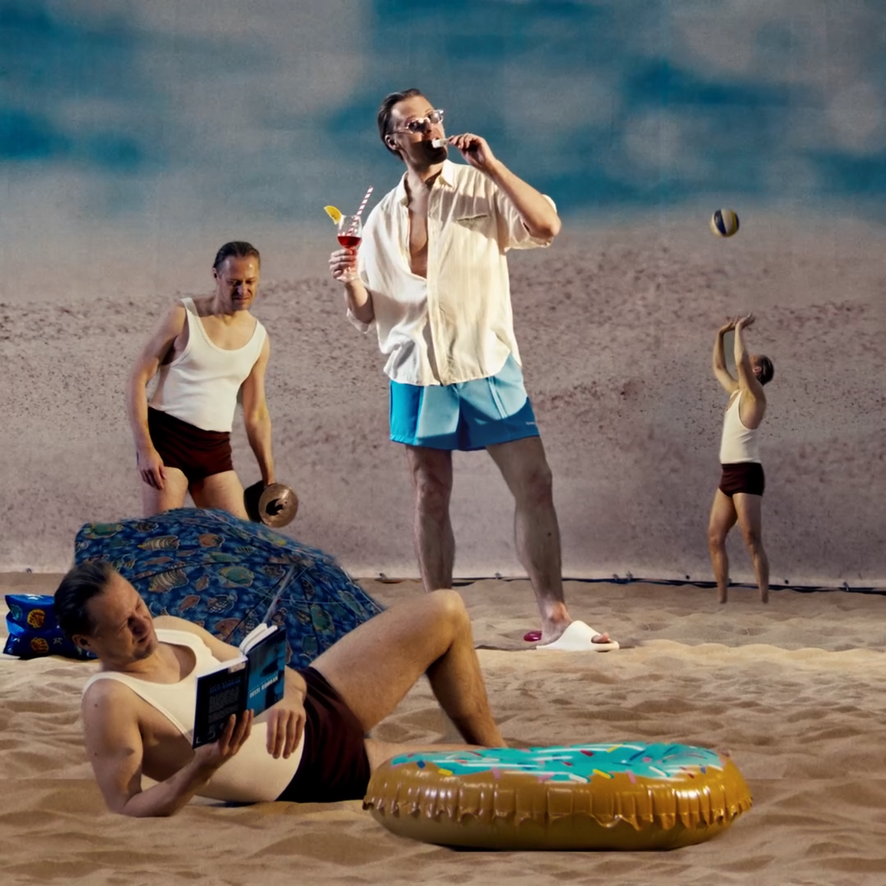 Group of people on a beach, some lying down and others standing. One person is holding a drink, and another is playing with a beach ball. The background features a calm sea and cloudy sky.