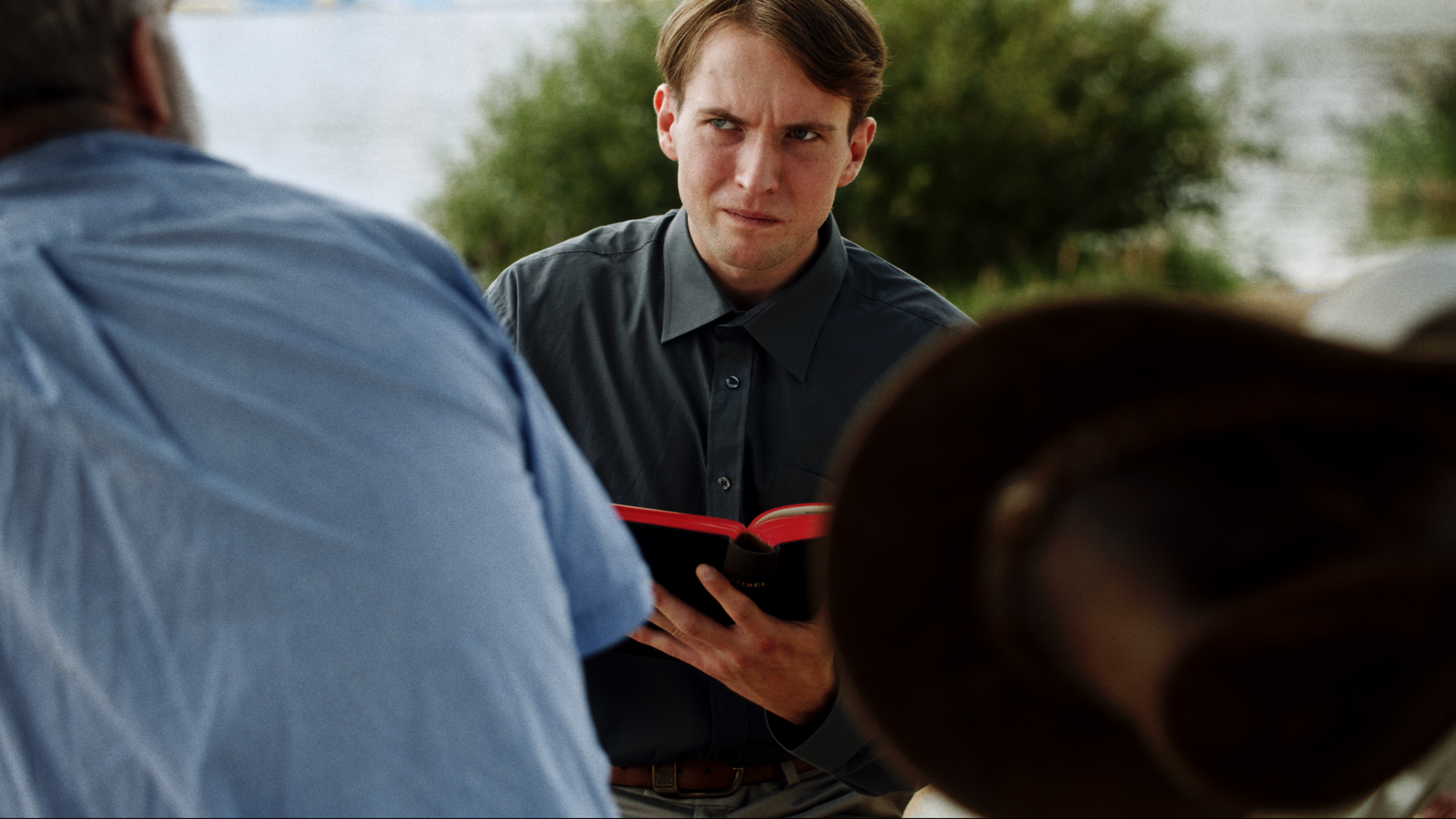 A young man with brown hair and a dark gray shirt, holding a small black book with a red edge, appears to be listening or speaking, with an outdoor background and a blurred person in the foreground.