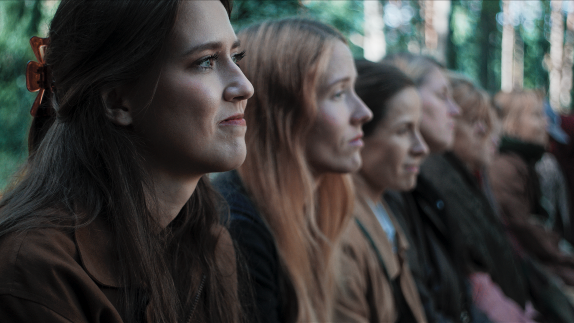 A group of women seated outdoors, looking to the right, with the background of trees, suggesting they are in a natural or park setting.