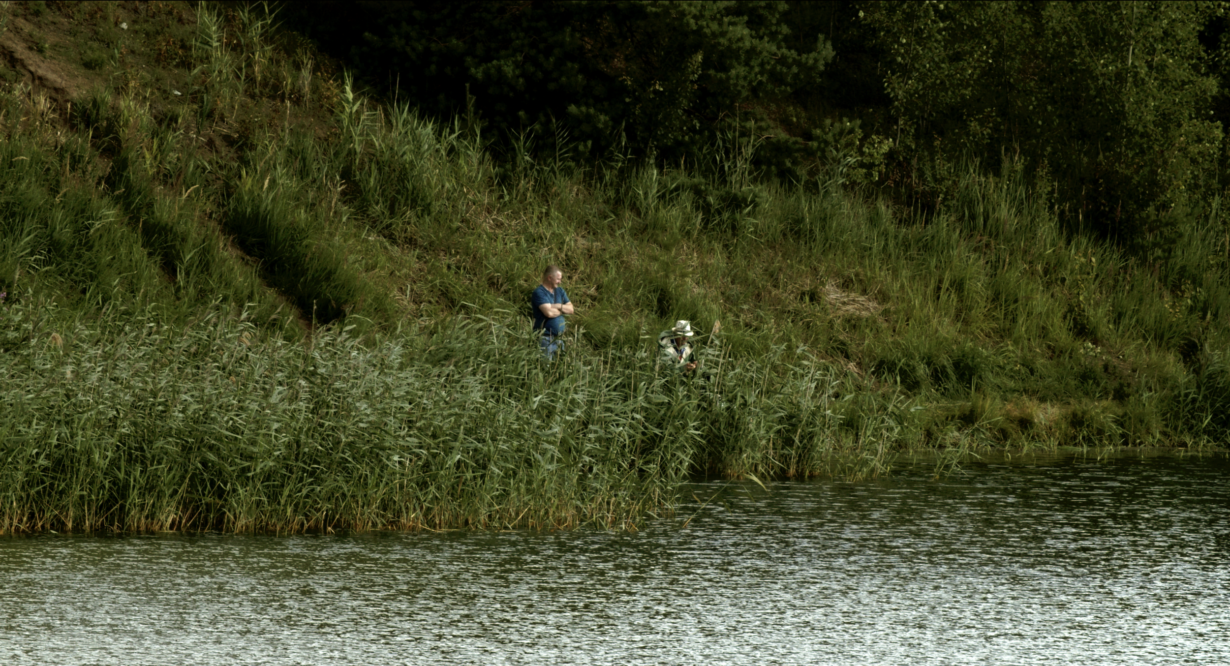 A man and a child standing on a grassy riverbank surrounded by tall green grass and trees, with water in the foreground.