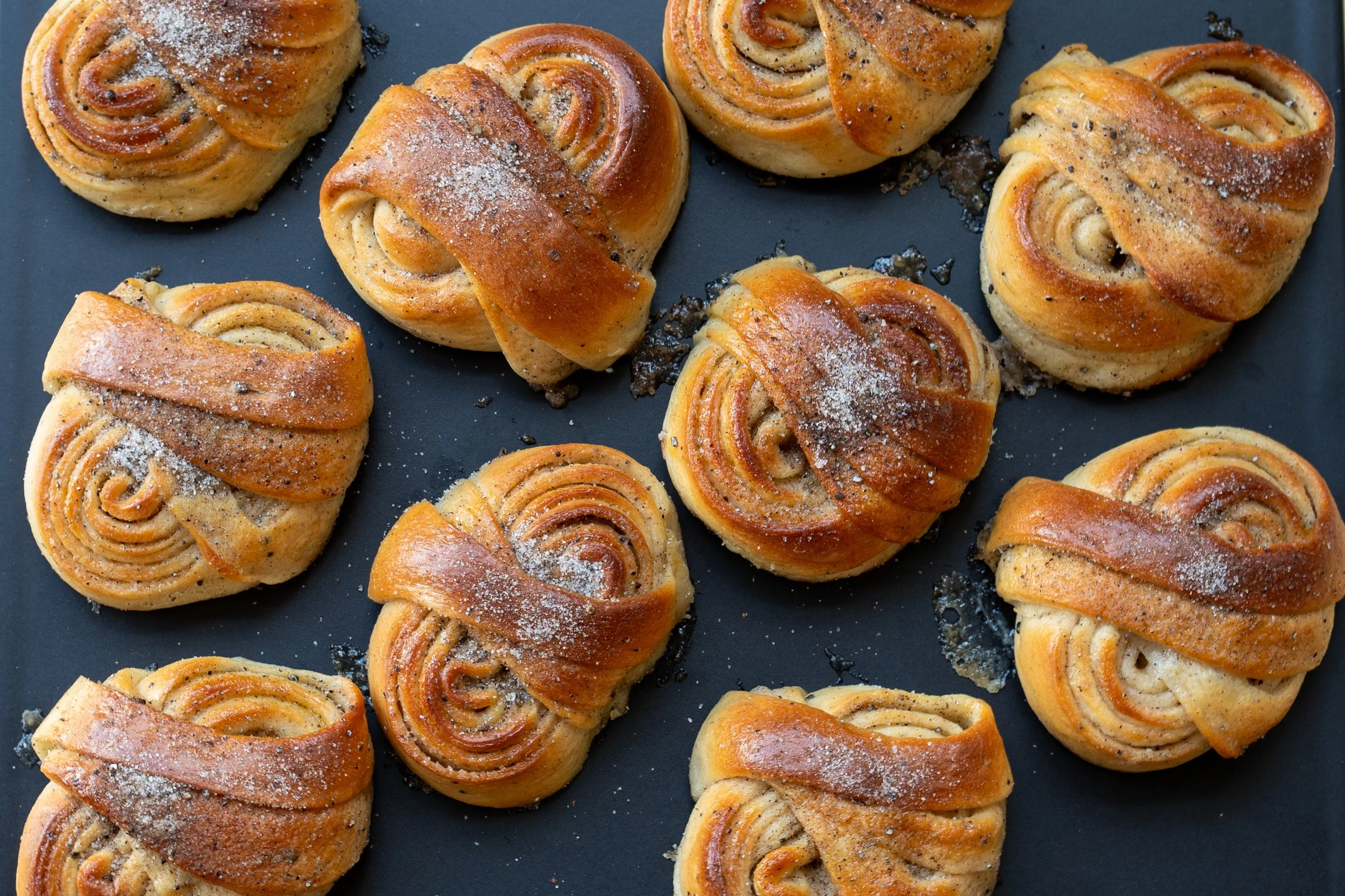 Several freshly baked cinnamon rolls on a dark baking sheet, topped with powdered sugar and black poppy seeds.