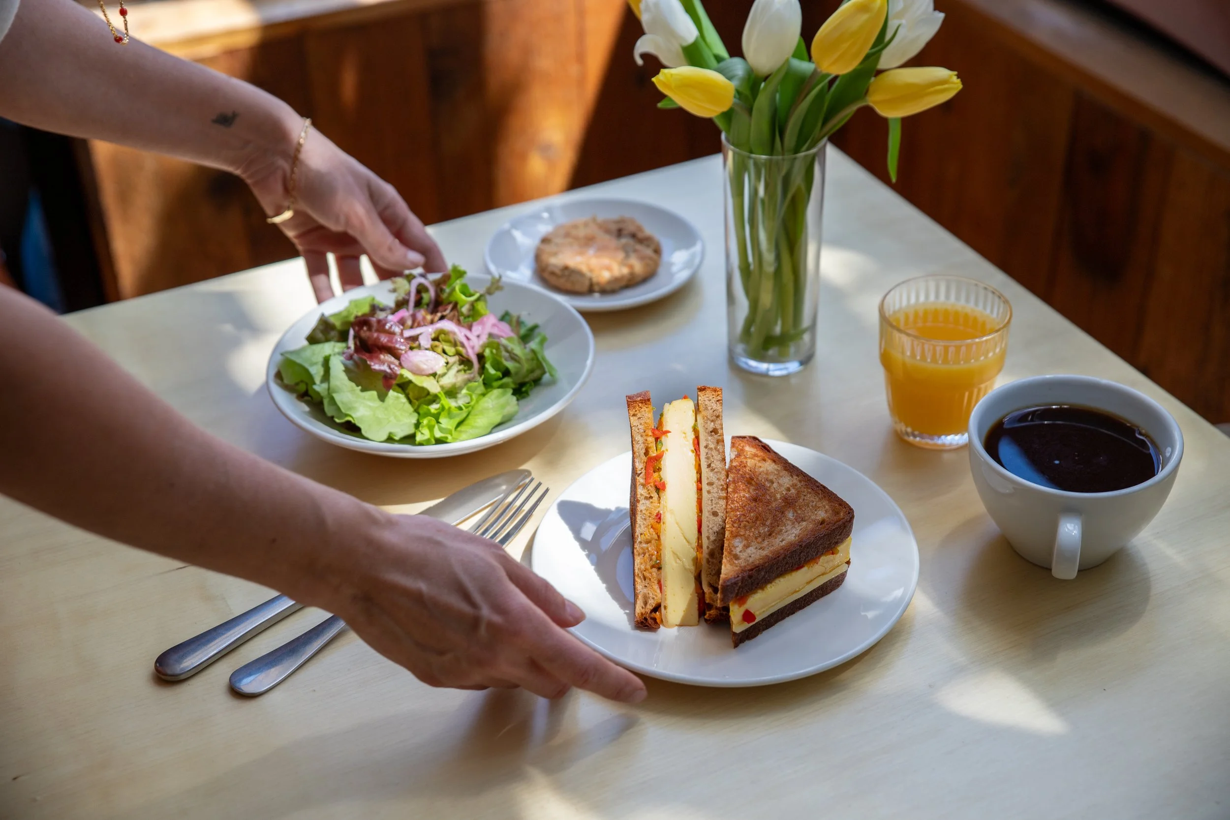 A breakfast table with a plate of club sandwich, a bowl of coffee, a glass of orange juice, a bowl with a cookie, a vase of yellow and white tulips, and a fresh salad. A person is placing the salad onto the plate.