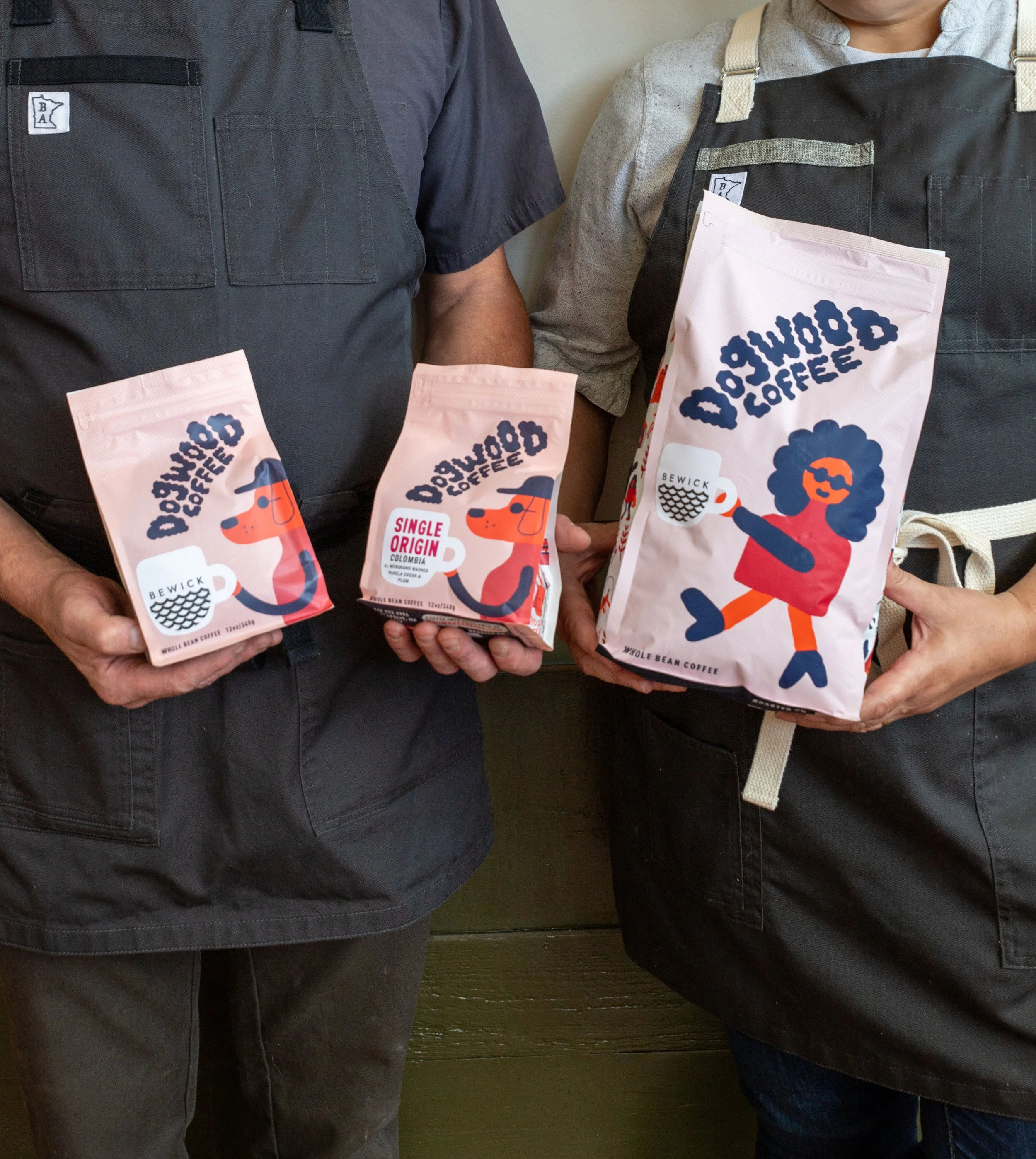 Two people wearing gray aprons, holding bags of coffee labeled 'Bewick Whole Bean Coffee,' standing side by side with their torsos visible.