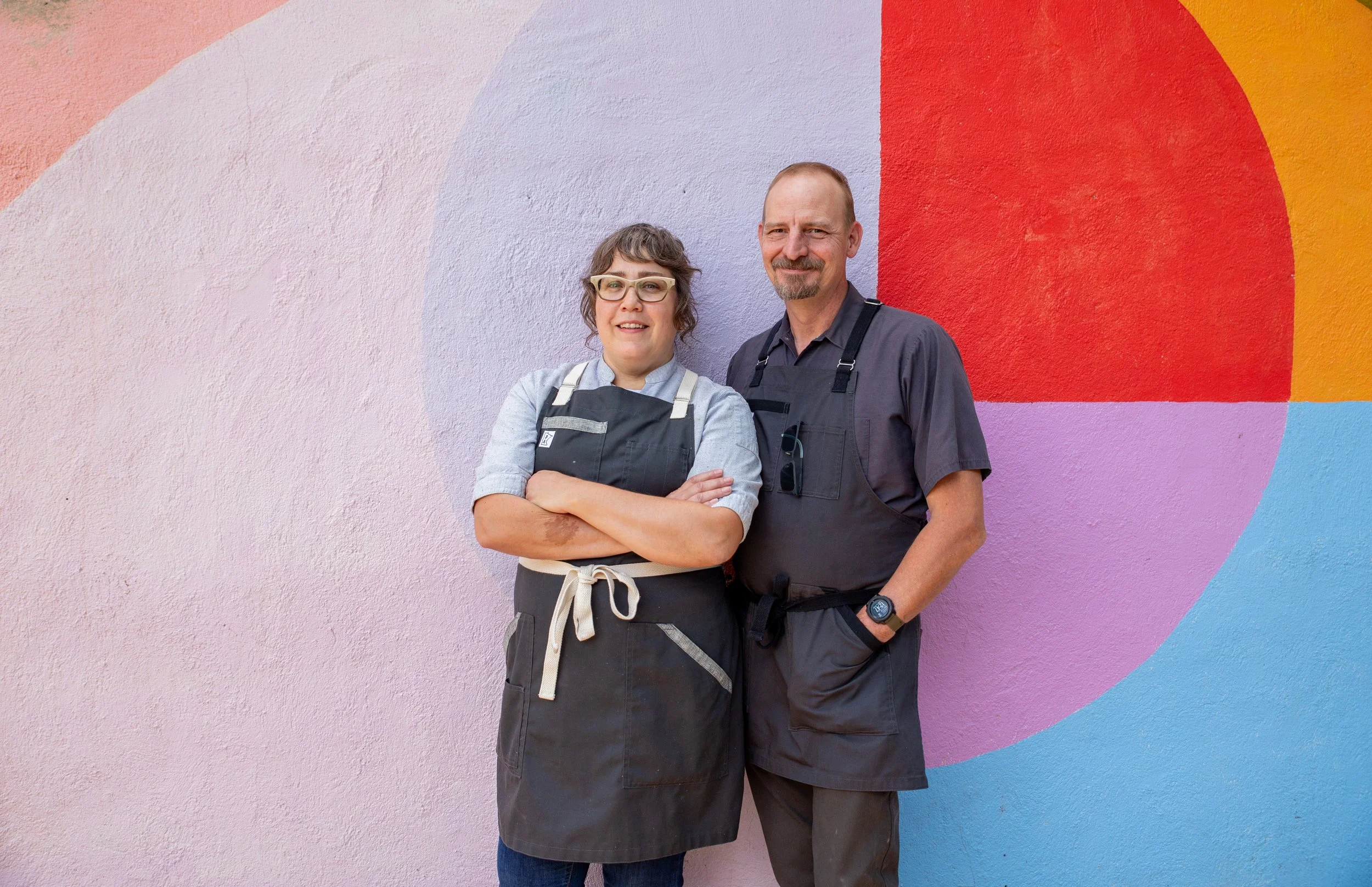 A woman with glasses and a man with a beard standing in front of a colorful mural wall, both wearing dark aprons.