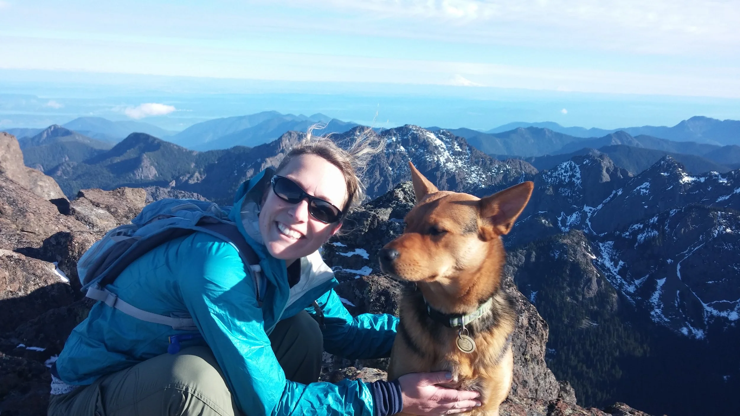 A woman smiling and wearing sunglasses, sitting on a mountain summit with her arm around a brown dog, with a panoramic view of mountain ridges and peaks behind them.