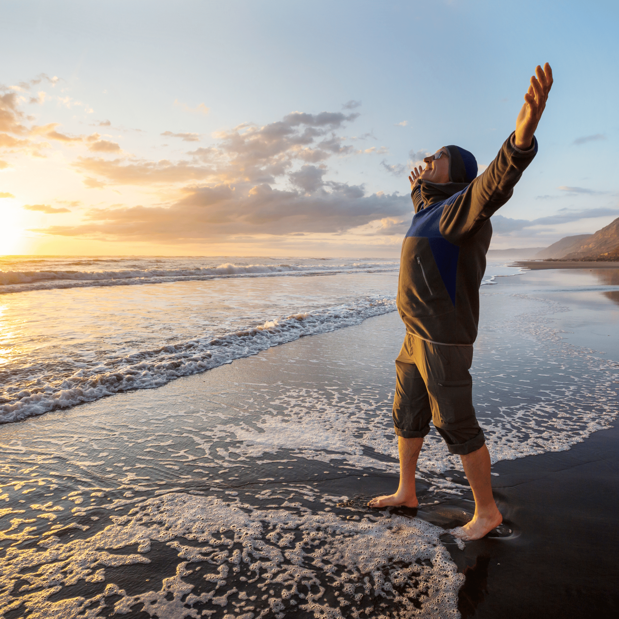 Un homme debout sur une plage au coucher du soleil, avec les bras levés et un sourire, face à l'océan.