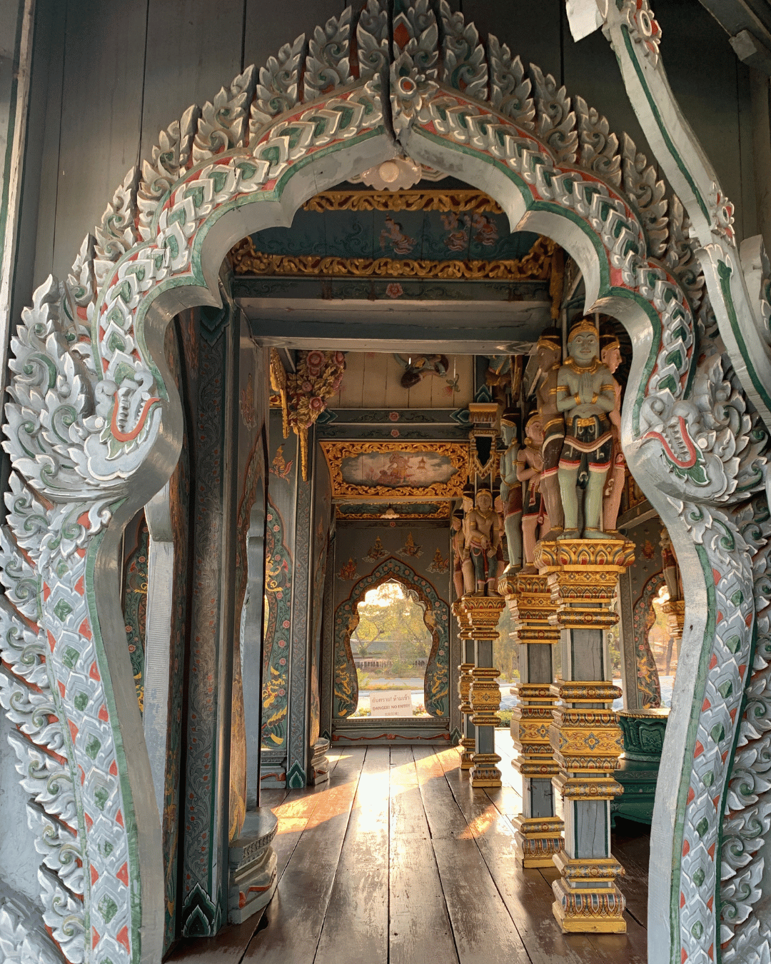 Intérieur d'un temple thaïlandais avec sculptures et décorations en bois peint, ornée de motifs de dragons, statues de divinités, et fenêtres laissant passer la lumière du soleil.
