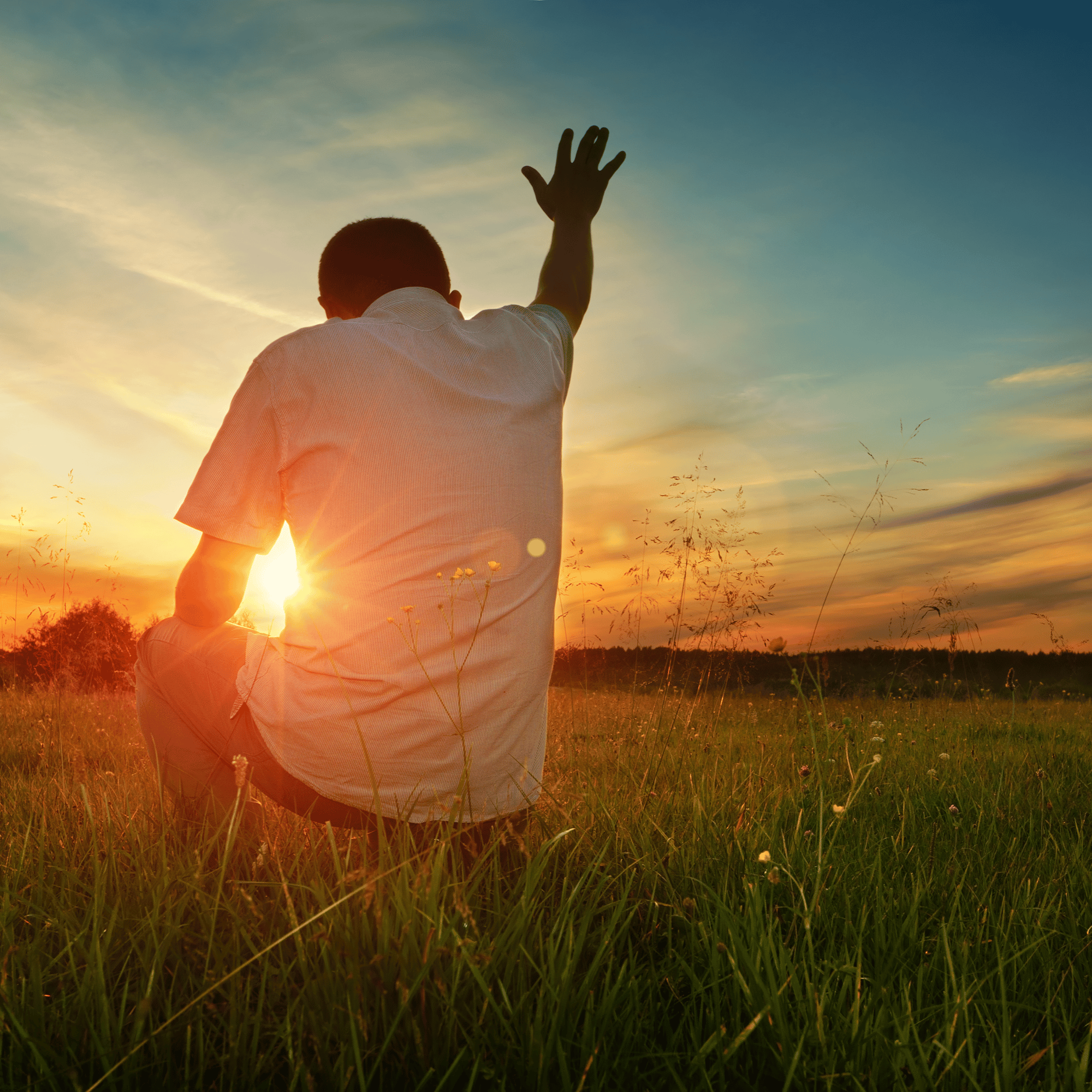 Un homme assis dans une prairie lors d'un coucher de soleil, levant la main en signe de salut ou de bénédiction.