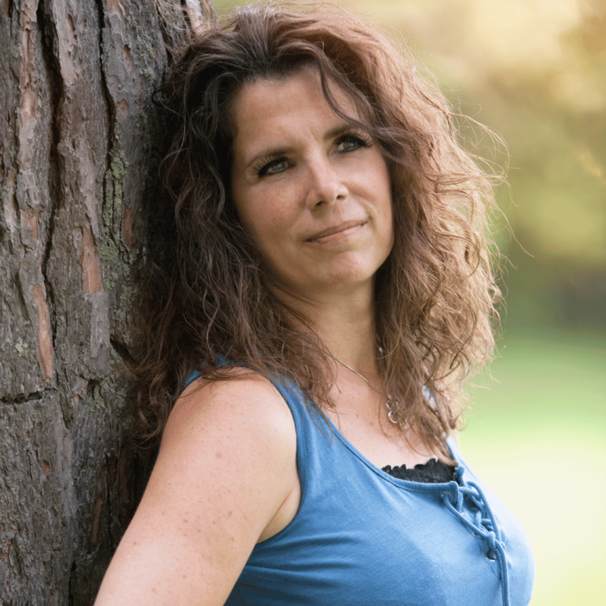 Une femme à cheveux bouclés bruns, portant un débardeur bleu, se tient contre un arbre dans un parc, regardant légèrement à gauche.