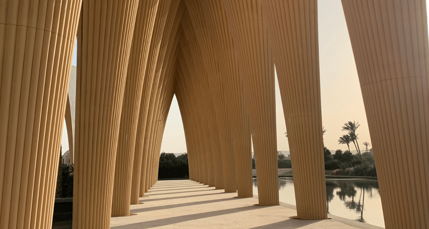Une passerelle architecturale en bois avec arches et colonnes en courbe, bordée par un miroir d'eau, avec des palmiers en arrière-plan au coucher du soleil.