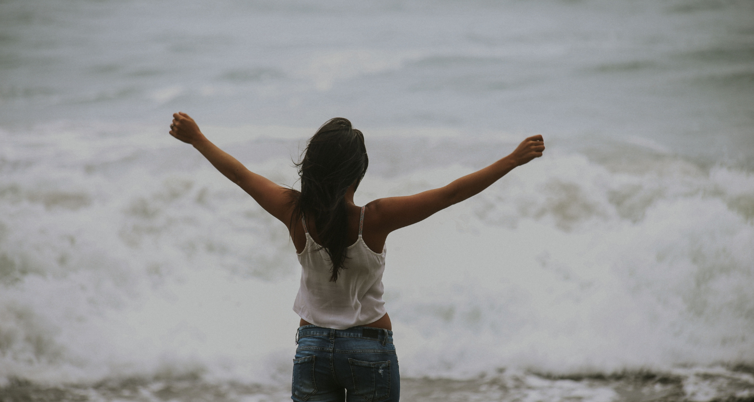 Une femme debout sur la plage avec les bras levés vers le ciel, face à la mer.