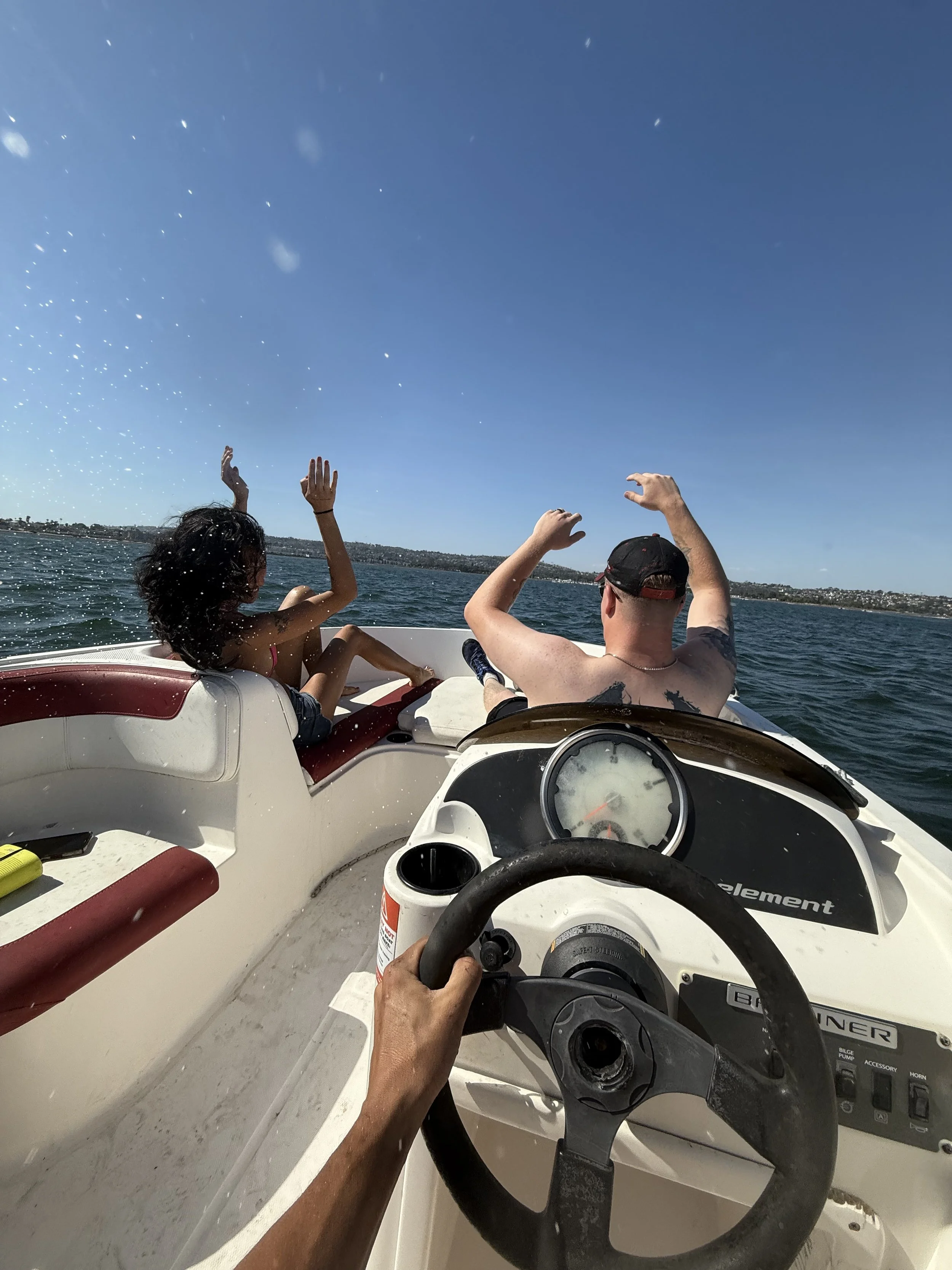 View from inside a boat showing two people, a young girl and an adult man, waving at the sky during a sunny day on the water, with a city or town visible in the distant background.