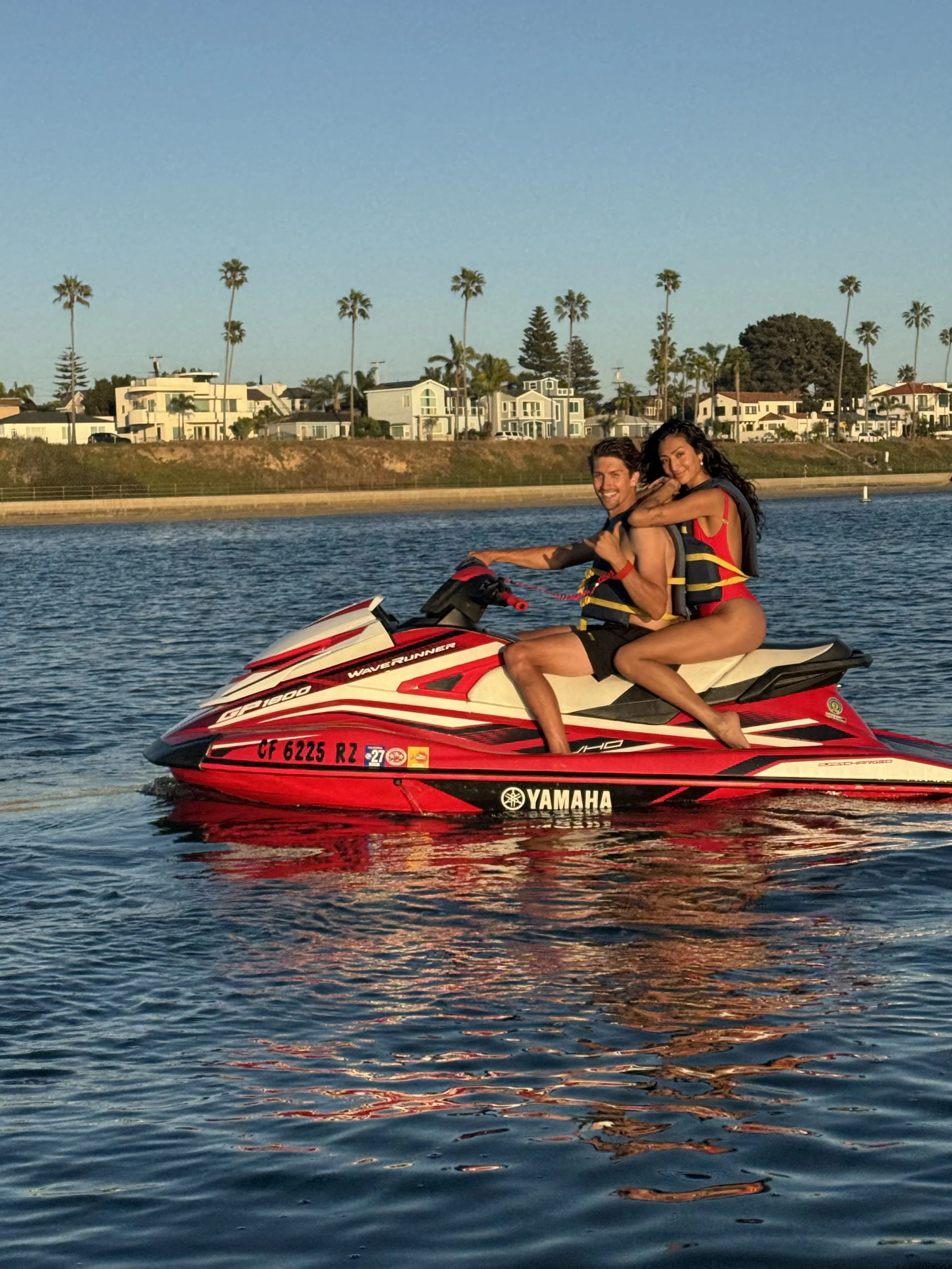 A man and woman riding a red Yamaha jet ski on a body of water with a background of houses and palm trees under a clear blue sky.