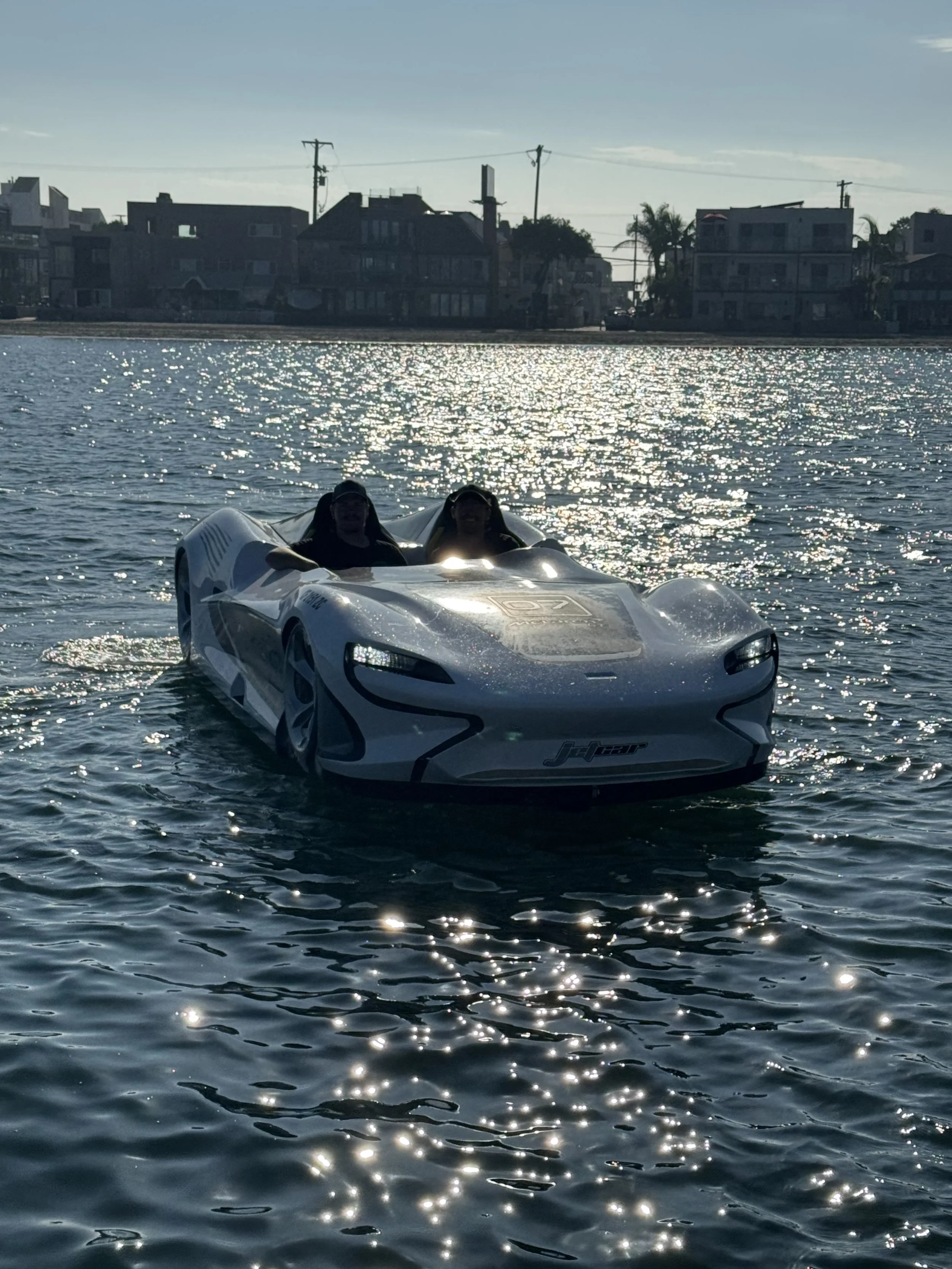 A futuristic white car used as a boat with two people inside, floating on water with a cityscape and houses in the background, reflected by the sunlight on the water.