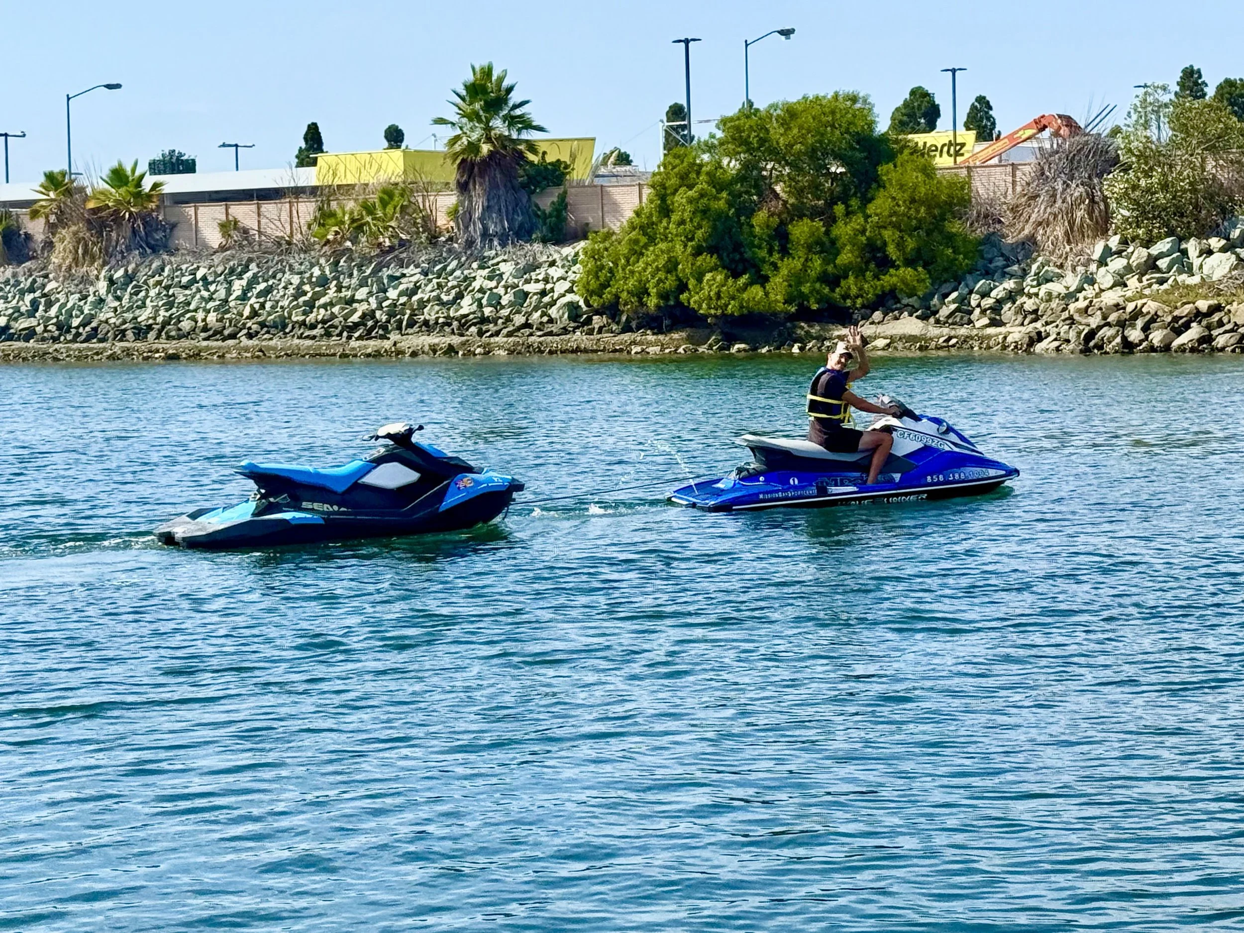 Person riding a blue jet ski on a body of water with trees and industrial buildings in the background.