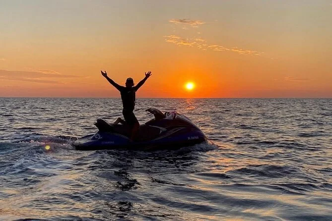Person on a jet ski with arms raised, riding on the water during a sunset over the ocean.