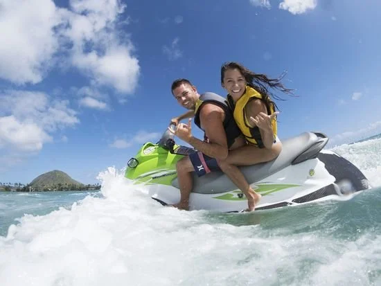 A smiling couple riding a jet ski on the water, with a clear blue sky and a small island in the background.